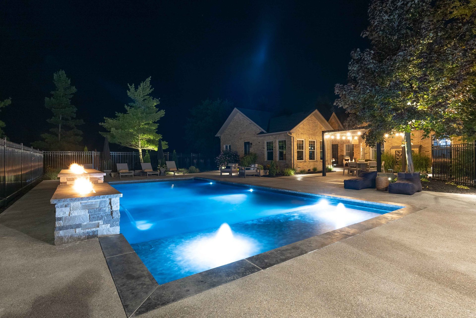 Illuminated backyard pool at night beside a patio and house with trees and stone landscaping