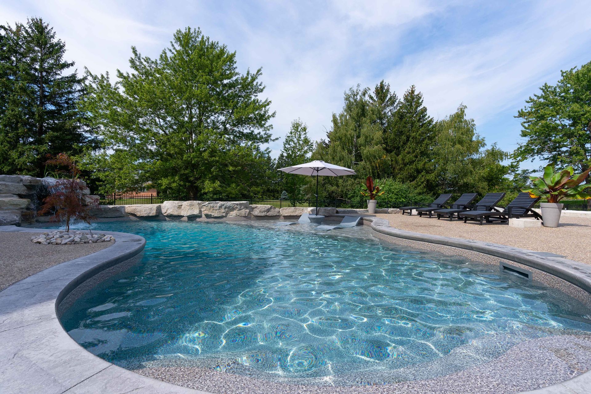 Backyard pool with turquoise water, stone coping, lounge chairs, and trees under a bright blue sky