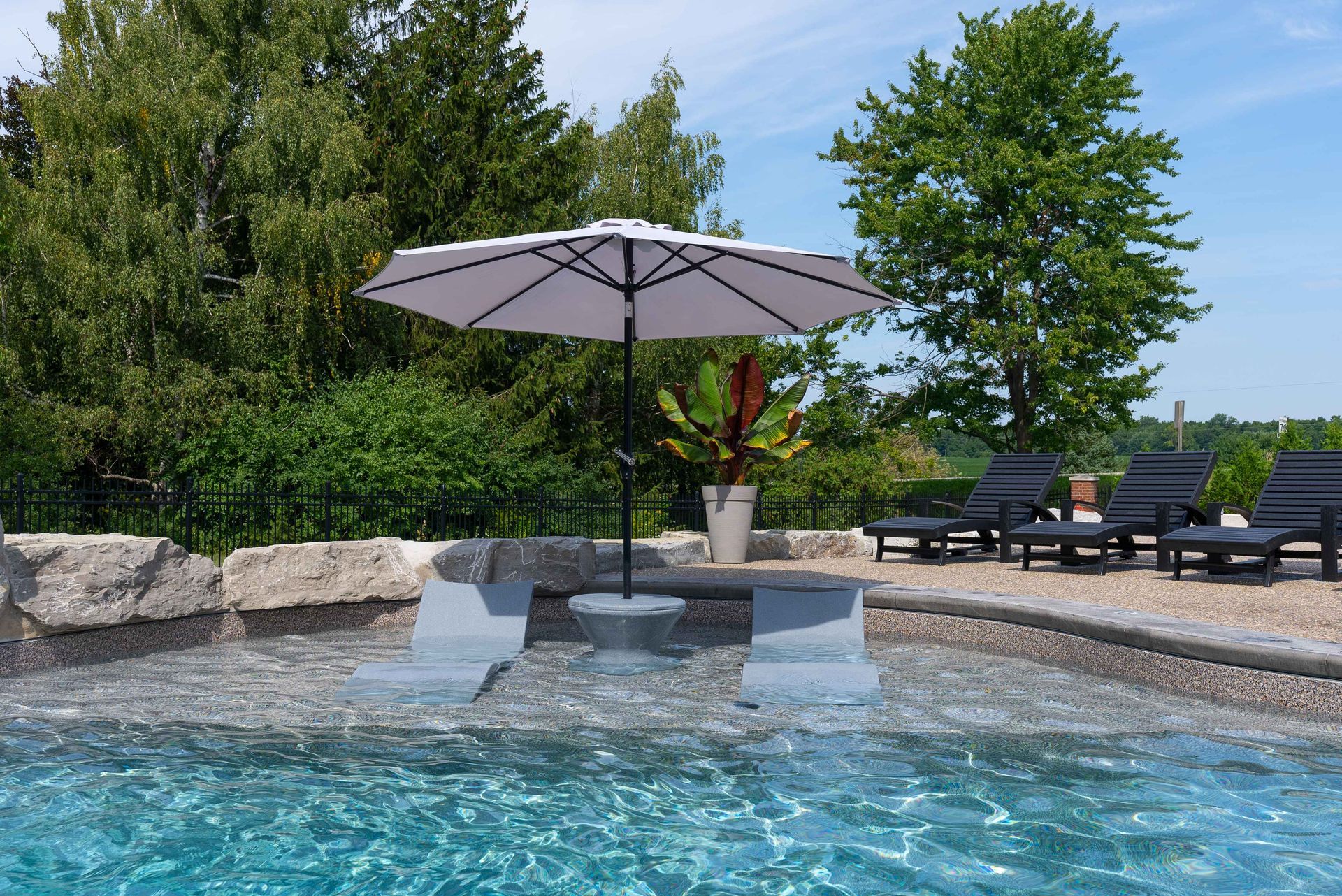 Poolside patio with umbrella-shaded table, lounge chairs, and turquoise water under a clear sky