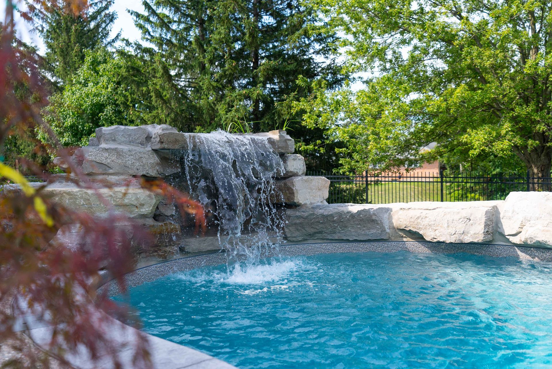 Backyard pool with a small rock waterfall and clear blue water, surrounded by trees and greenery
