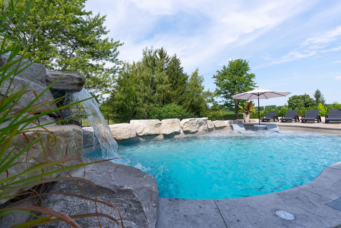 Backyard pool with waterfall, blue water, stone edges, and patio umbrella under a sunny sky