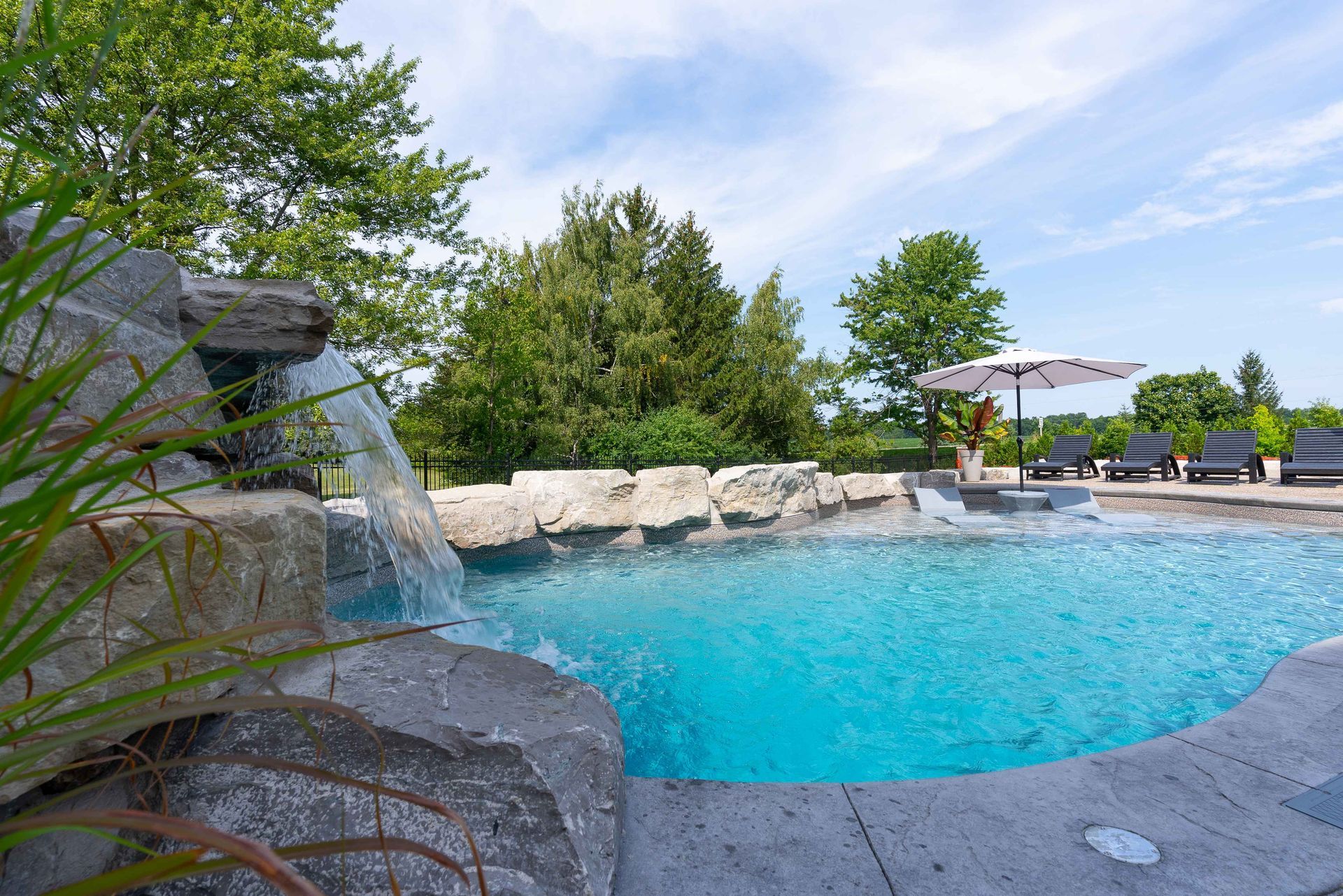 Outdoor pool with a waterfall, stone edge, and lounge chairs under umbrellas on a sunny day