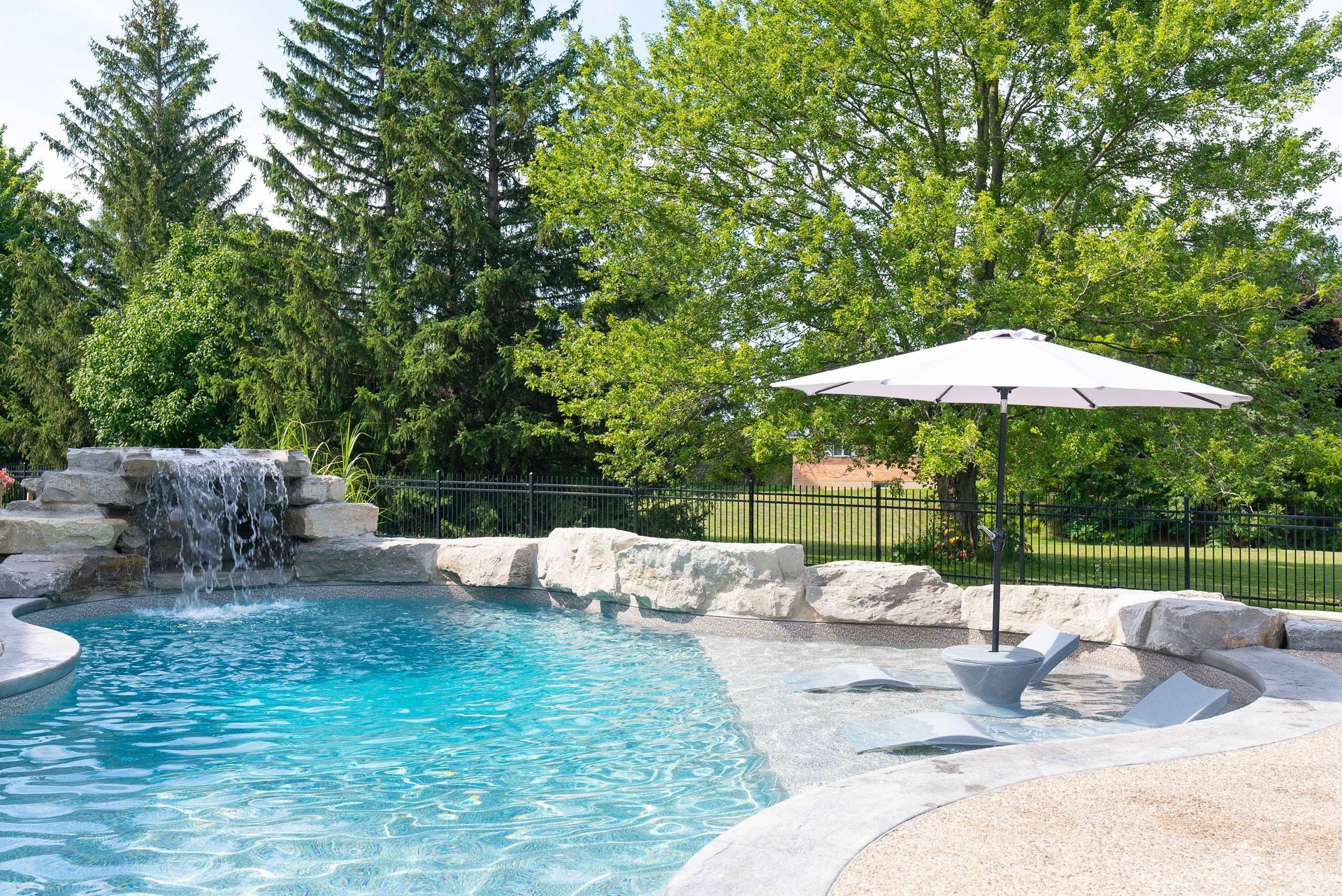 Backyard pool with waterfall, white umbrella, lounge chairs, and lush green trees