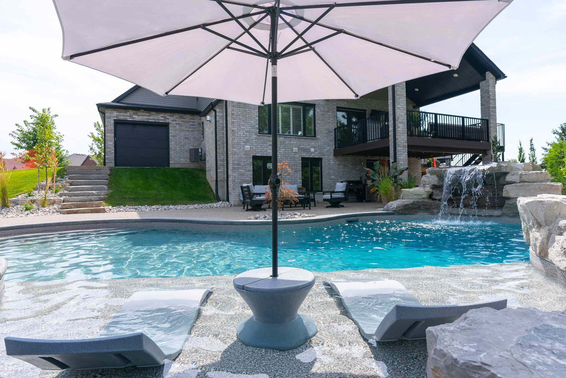 Backyard pool with large patio umbrella in front of a stone house and lounge chairs