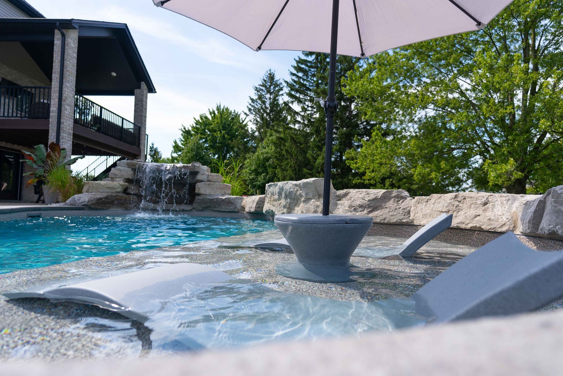 Poolside lounge chairs half-submerged in a blue spa beneath a white umbrella, with rocks and trees nearby