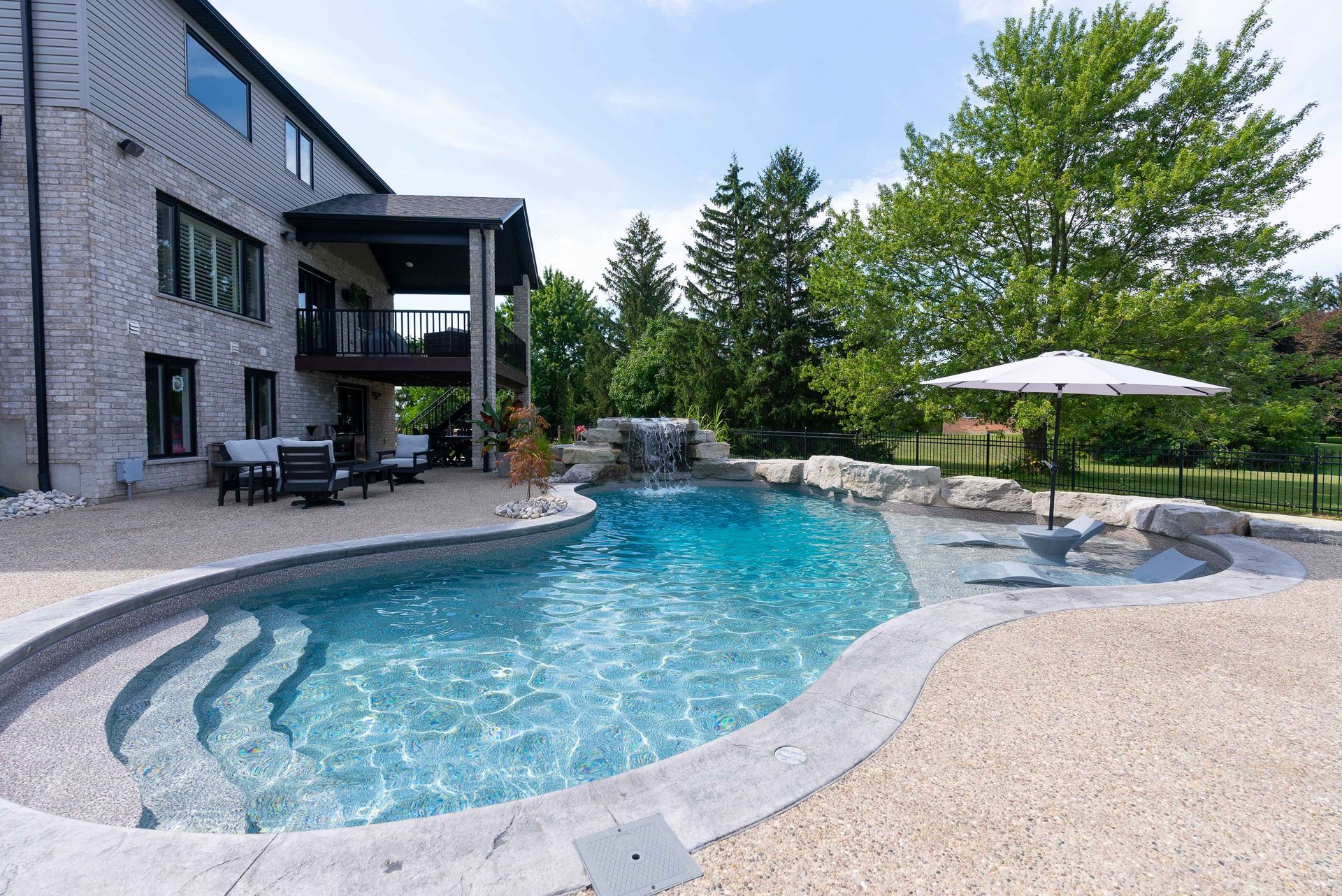 Modern backyard patio with a kidney-shaped swimming pool, stone deck, lounge chairs, and trees.