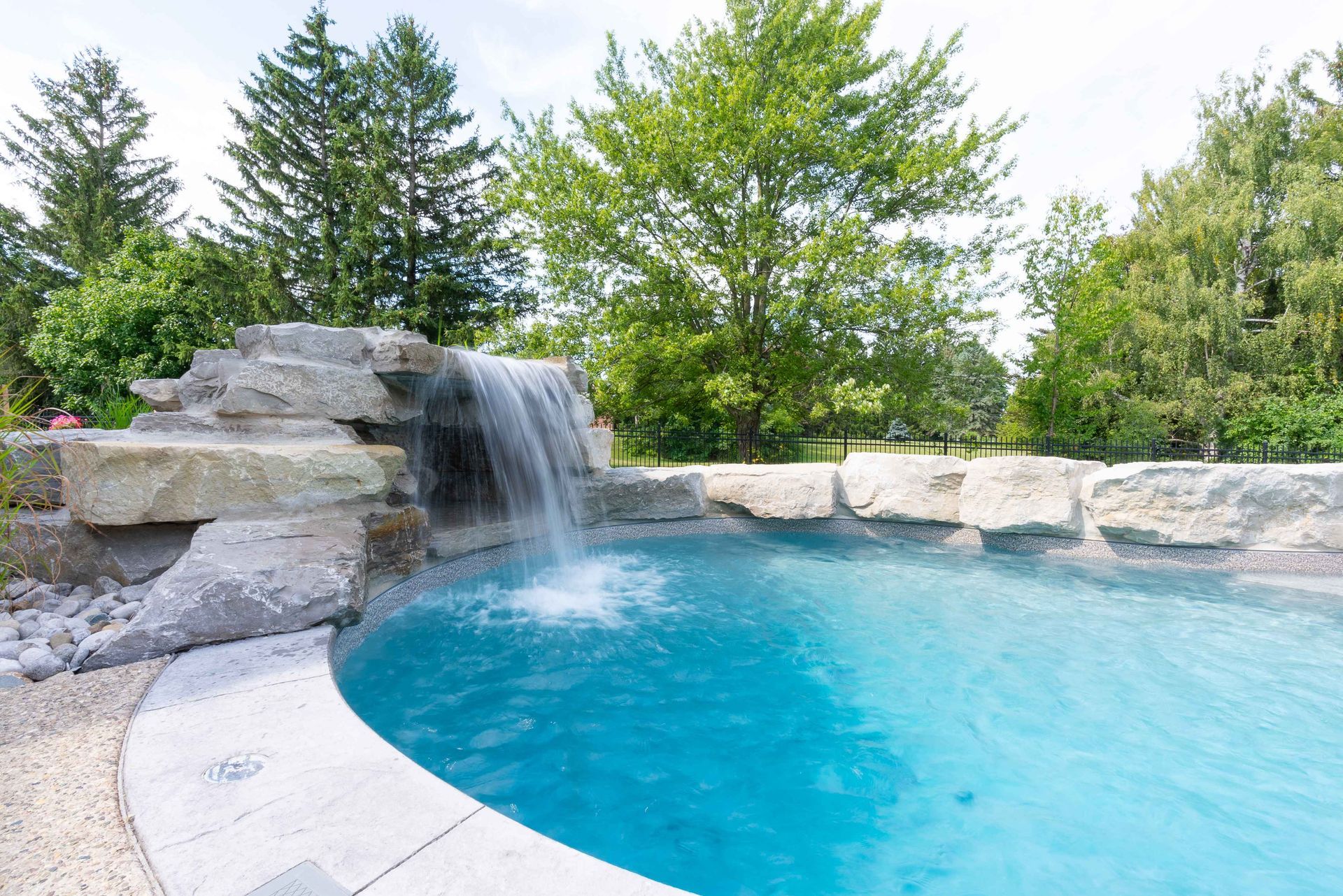 Blue backyard pool with a small rock waterfall and trees in the background