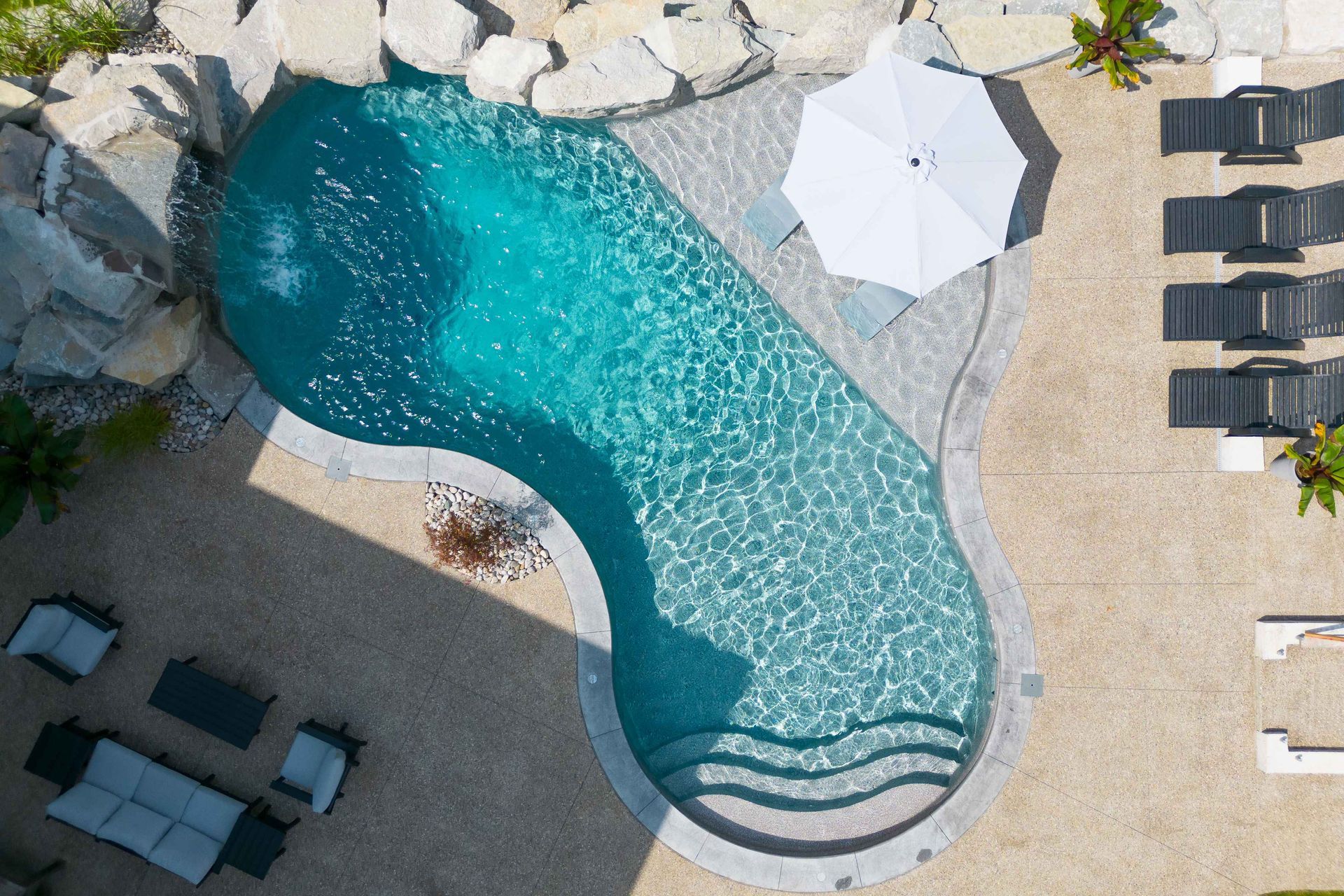 Aerial view of a curvy blue pool beside lounge chairs and a white umbrella on a stone patio.