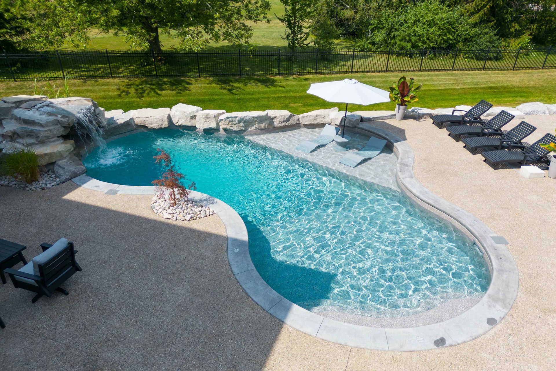 Backyard pool with curved blue water, stone deck, lounge chairs, and umbrella overlooking a green lawn
