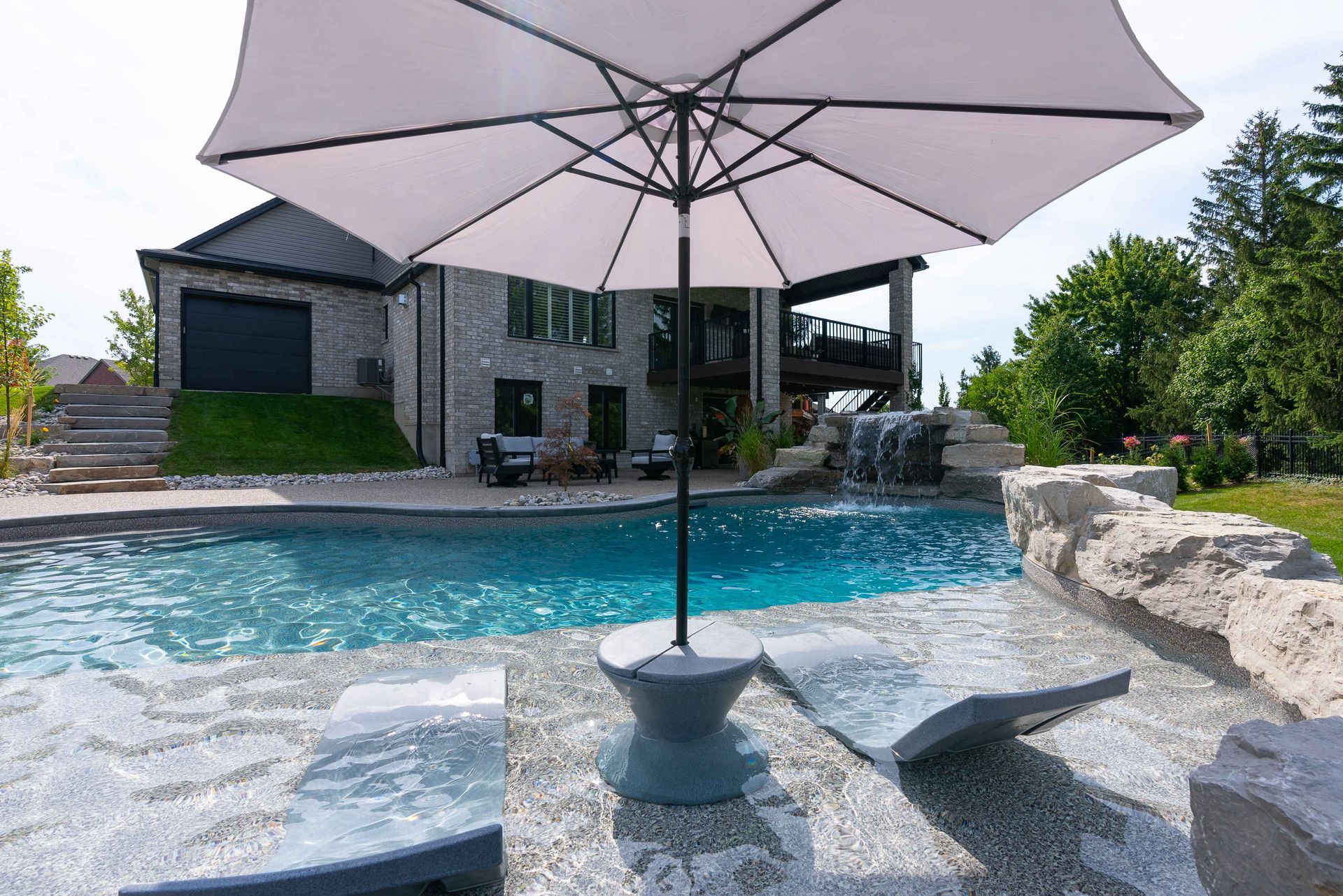 Backyard pool with patio umbrella and stone lounge chairs near a modern house