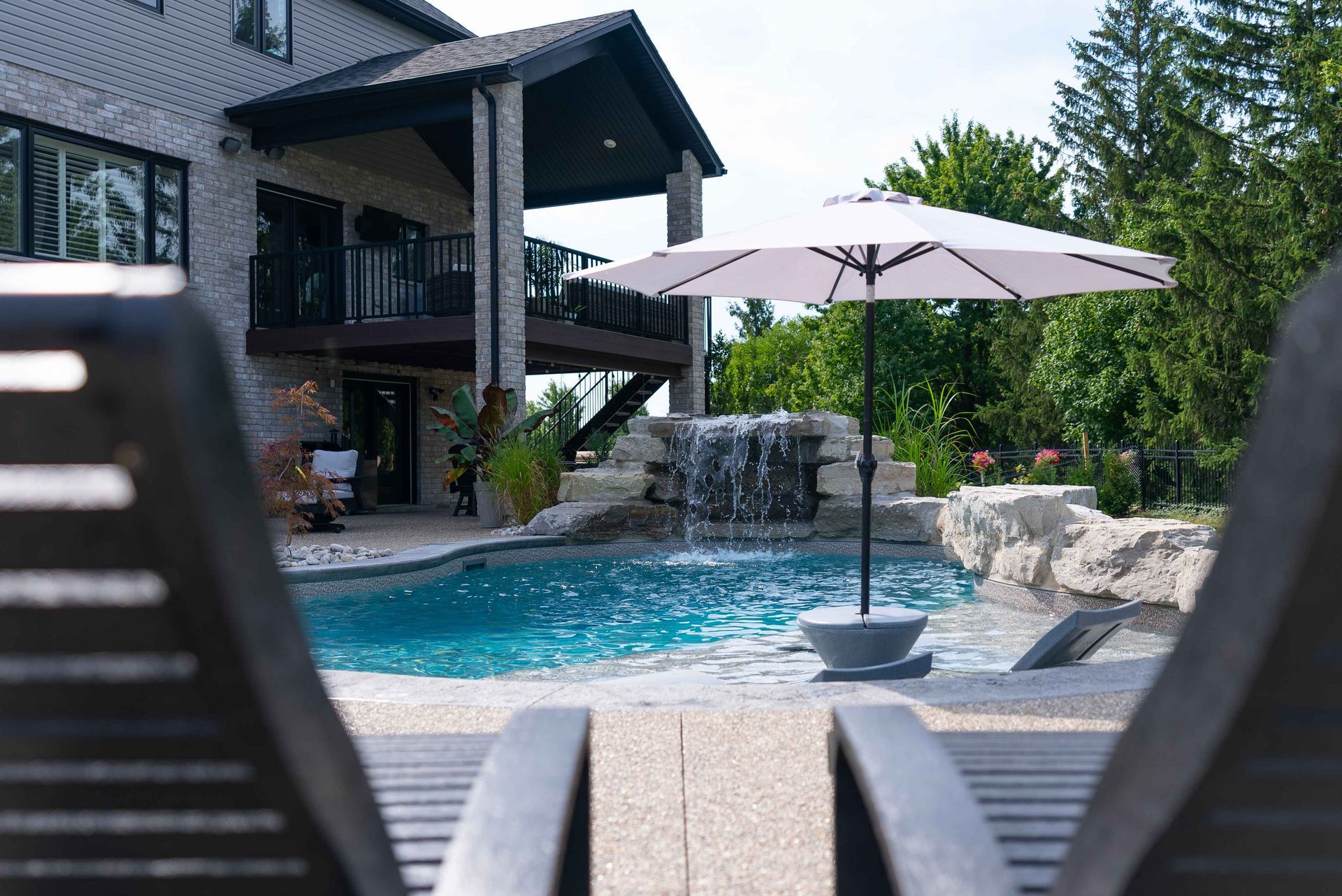 Backyard pool with waterfall, patio umbrella, and modern house, framed by lounge chairs