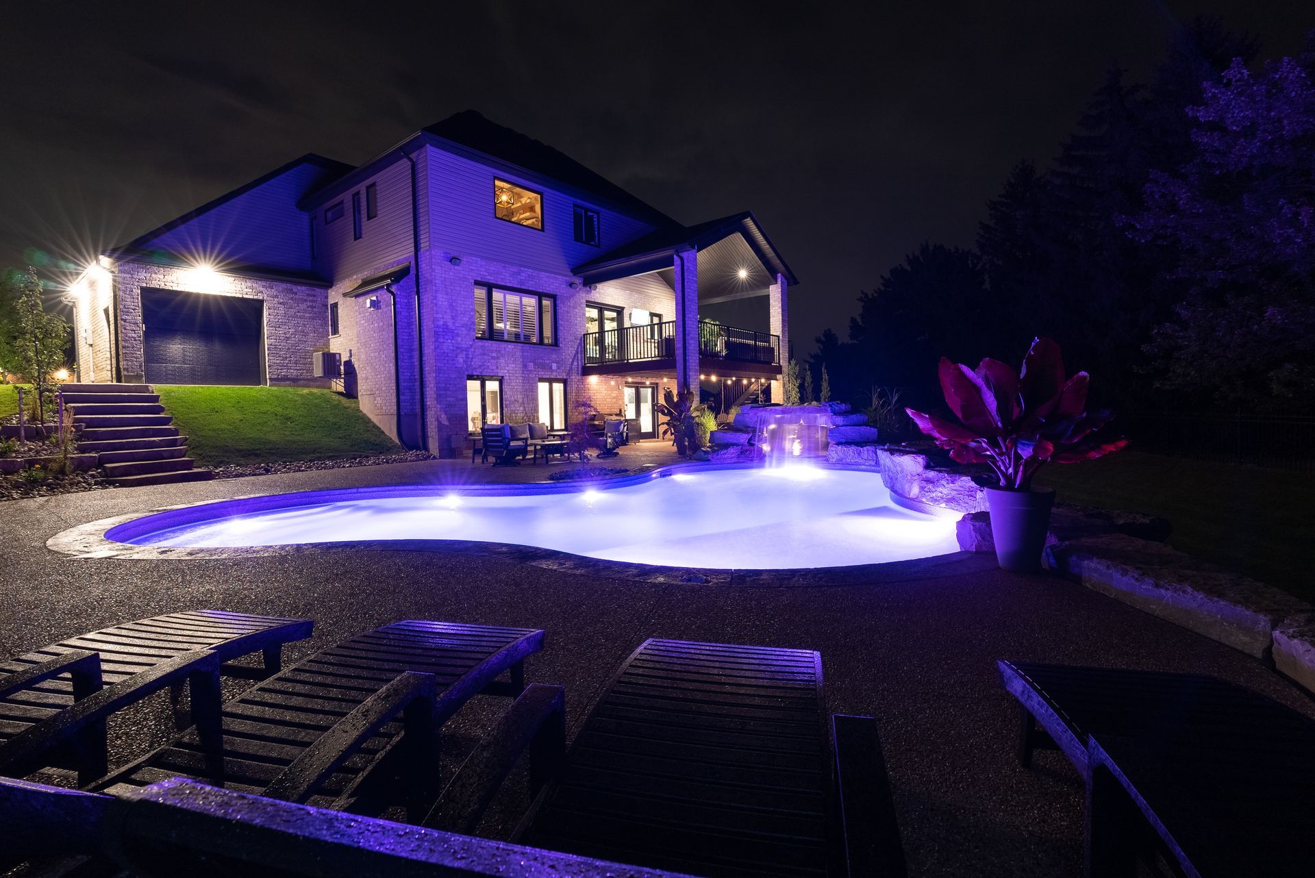 Nighttime view of a lit house with a glowing blue pool and patio furniture in the foreground