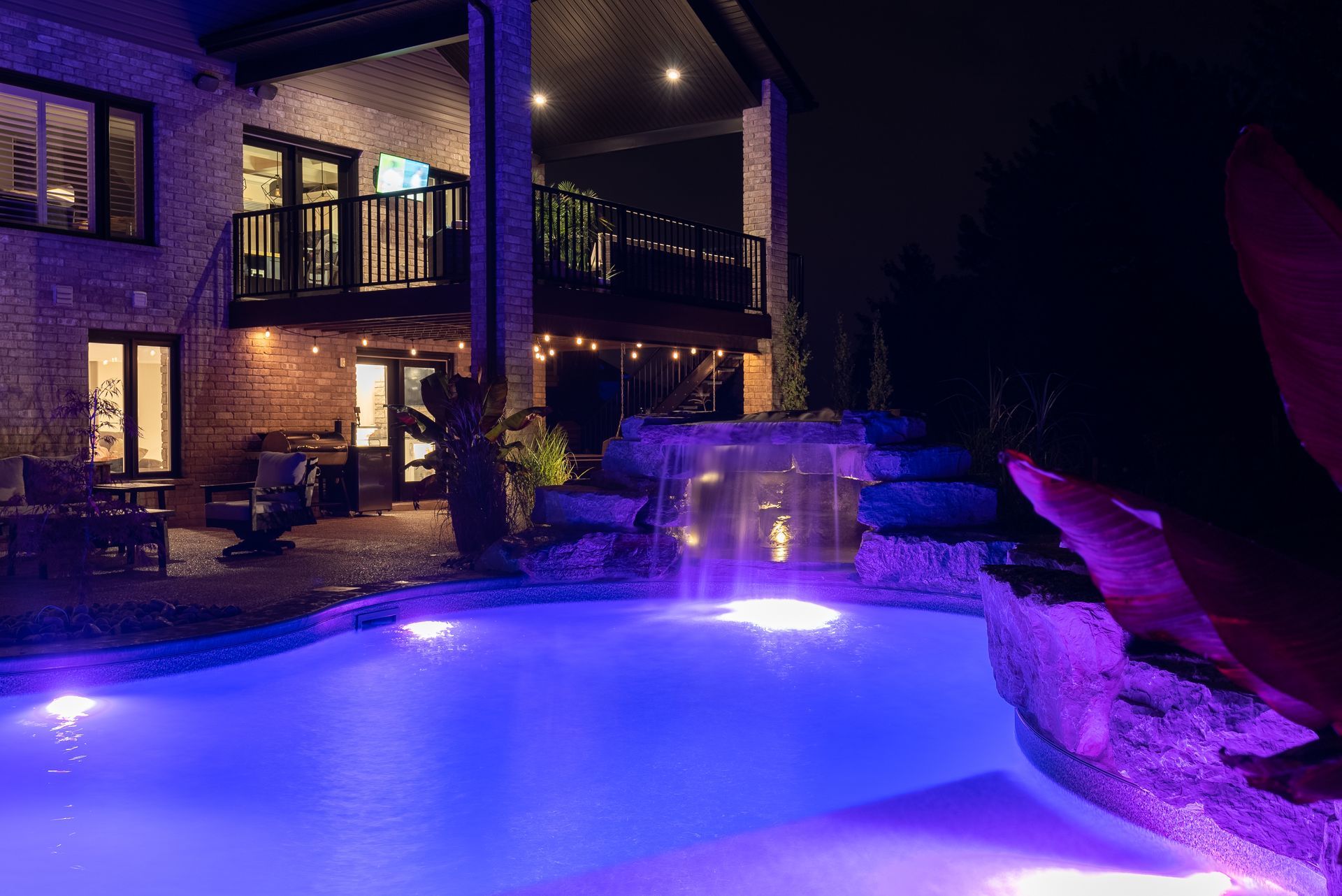 Purple-lit backyard pool beside a brick house and patio at night