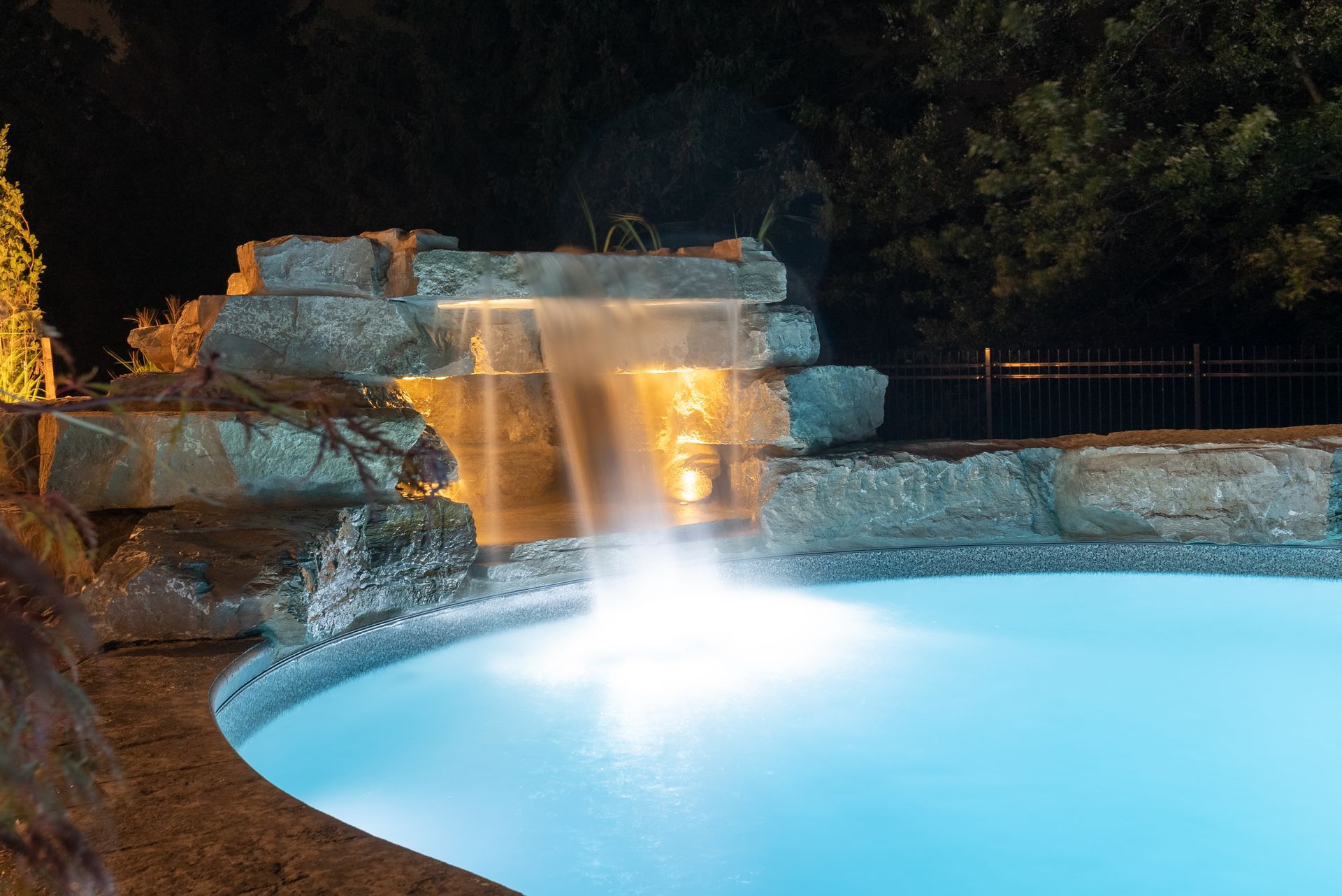 Illuminated backyard pool with a rock waterfall and glowing blue water at night