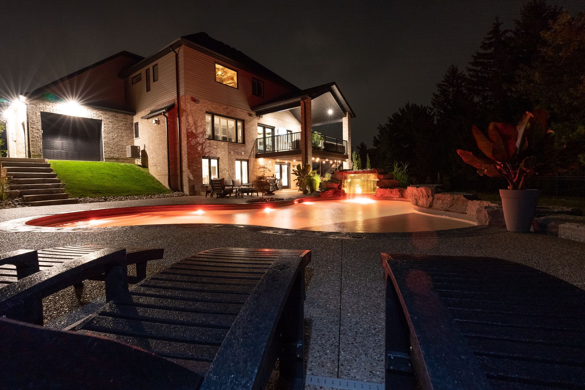 Nighttime view of a modern house with lit windows, red garden lights, and two lounge chairs in the foreground