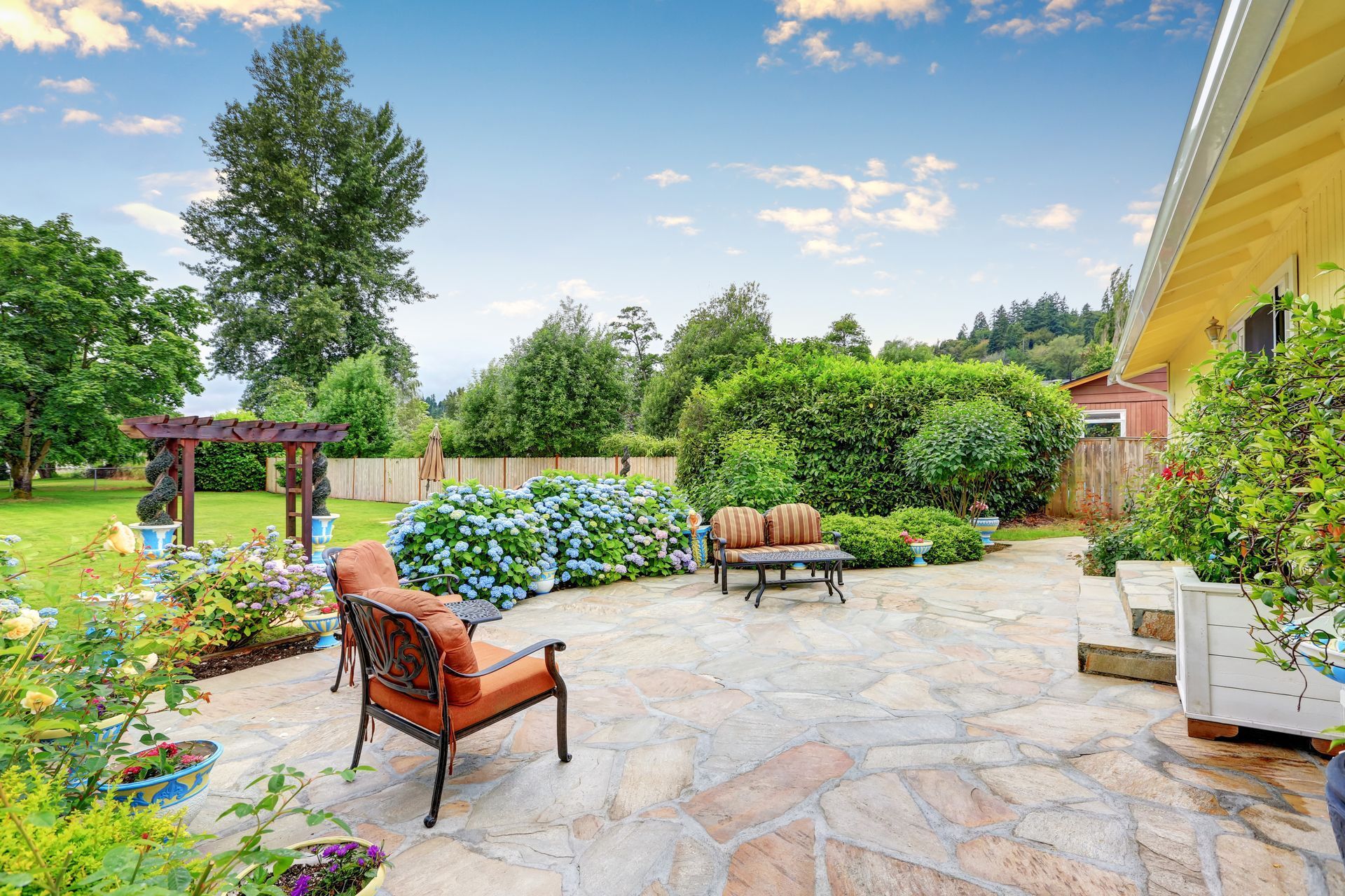A flagstone patio featuring orange chairs and a small table, surrounded by lush green landscaping and trees under a blue sky.