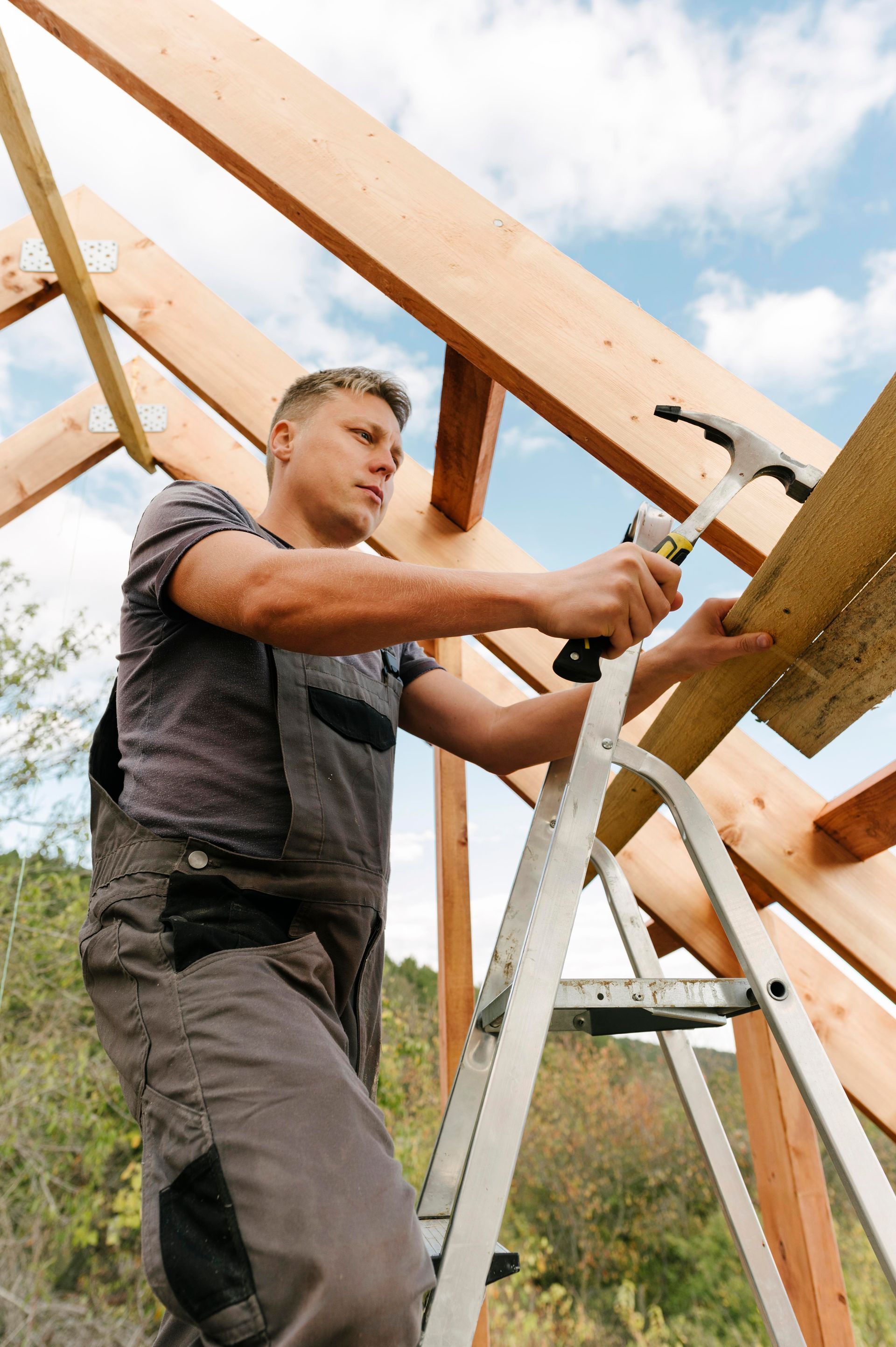 A person standing on a ladder hammers a wooden beam into place on the framing of an outdoor structure.