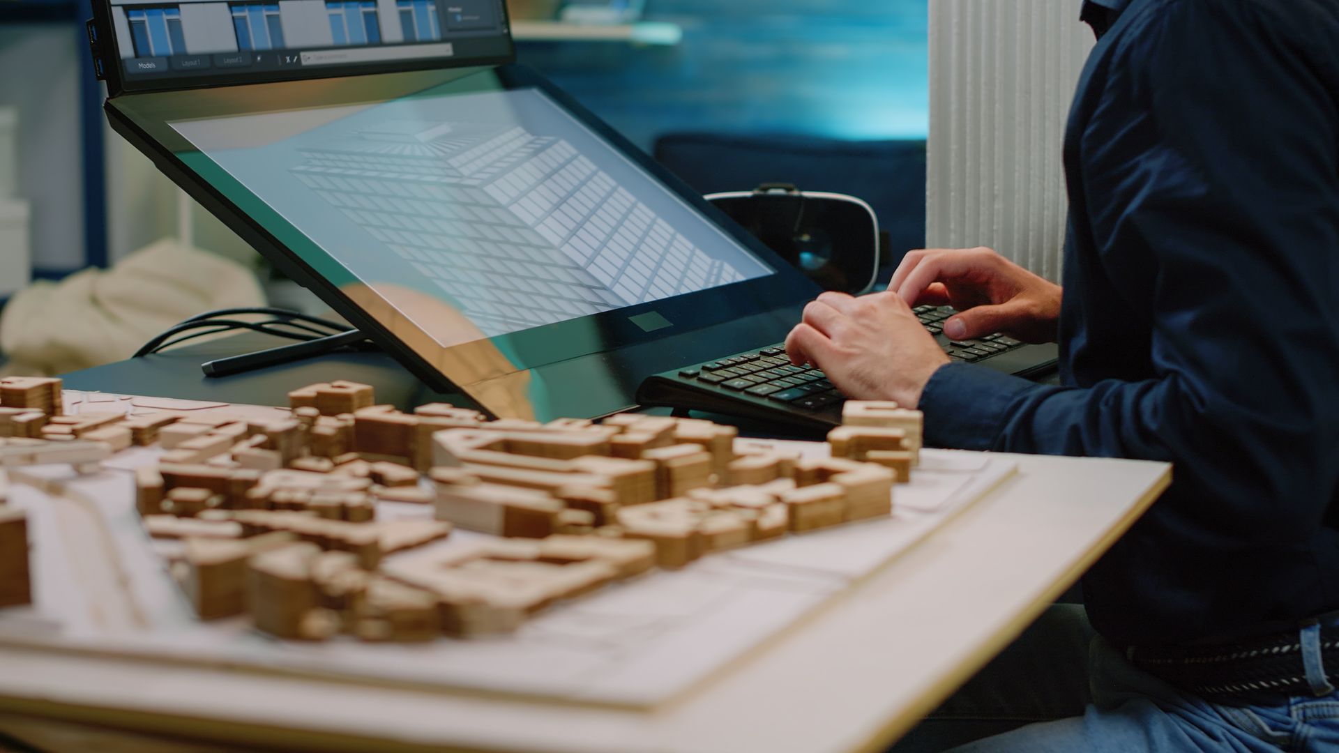 An architect working at a desk with a computer monitor displaying a building design next to a miniature urban model.