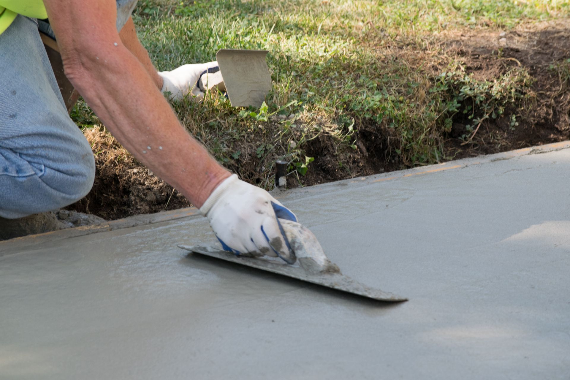 A person wearing a glove smooths fresh wet concrete on a sidewalk with a metal trowel outdoors.
