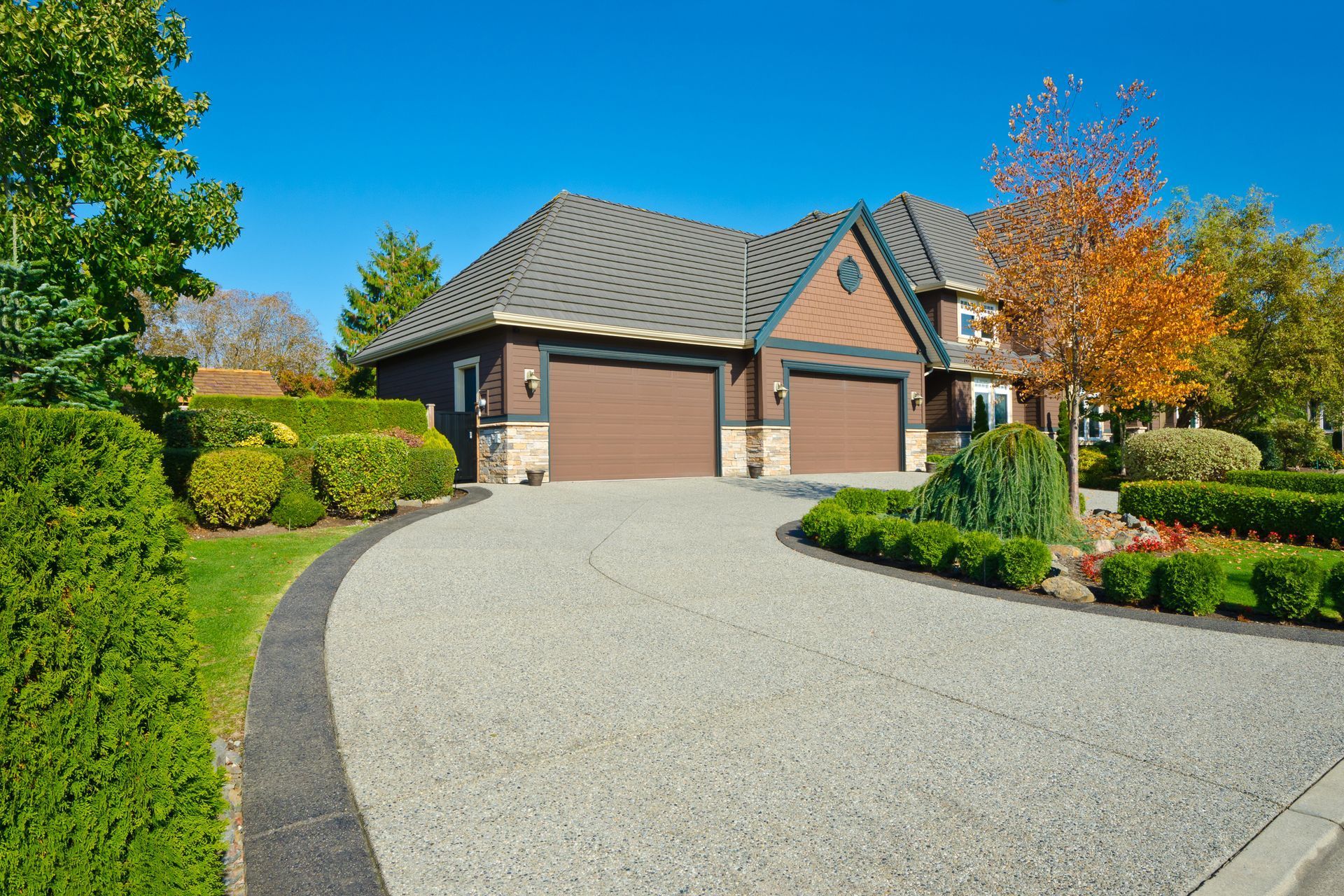 A suburban house with a two-car garage and a curved driveway, surrounded by green lawns and autumn trees under a blue sky.
