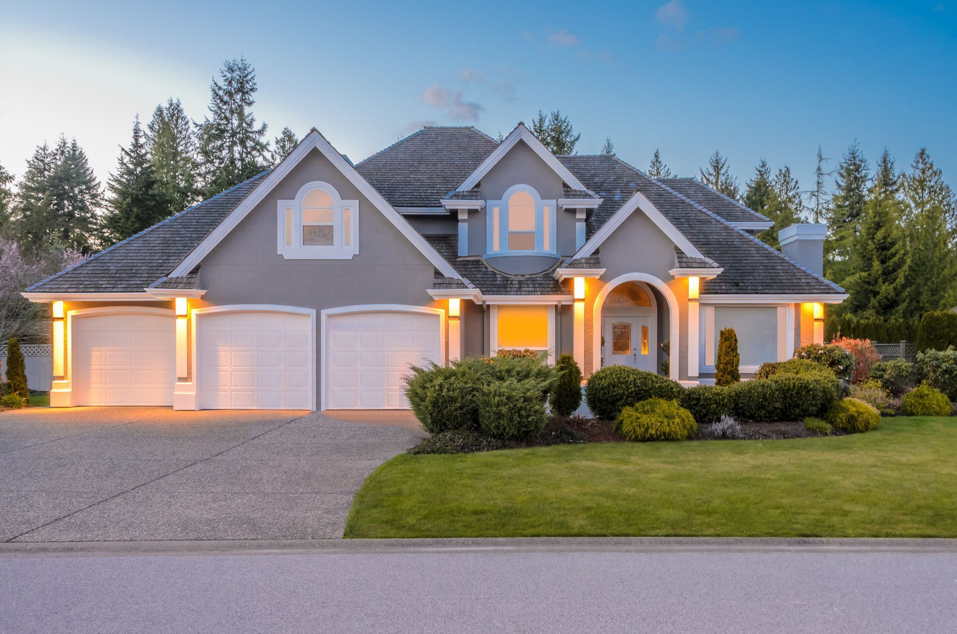 A gray, two-story house with a three-car garage, manicured lawn, and evergreen trees under a twilight sky.