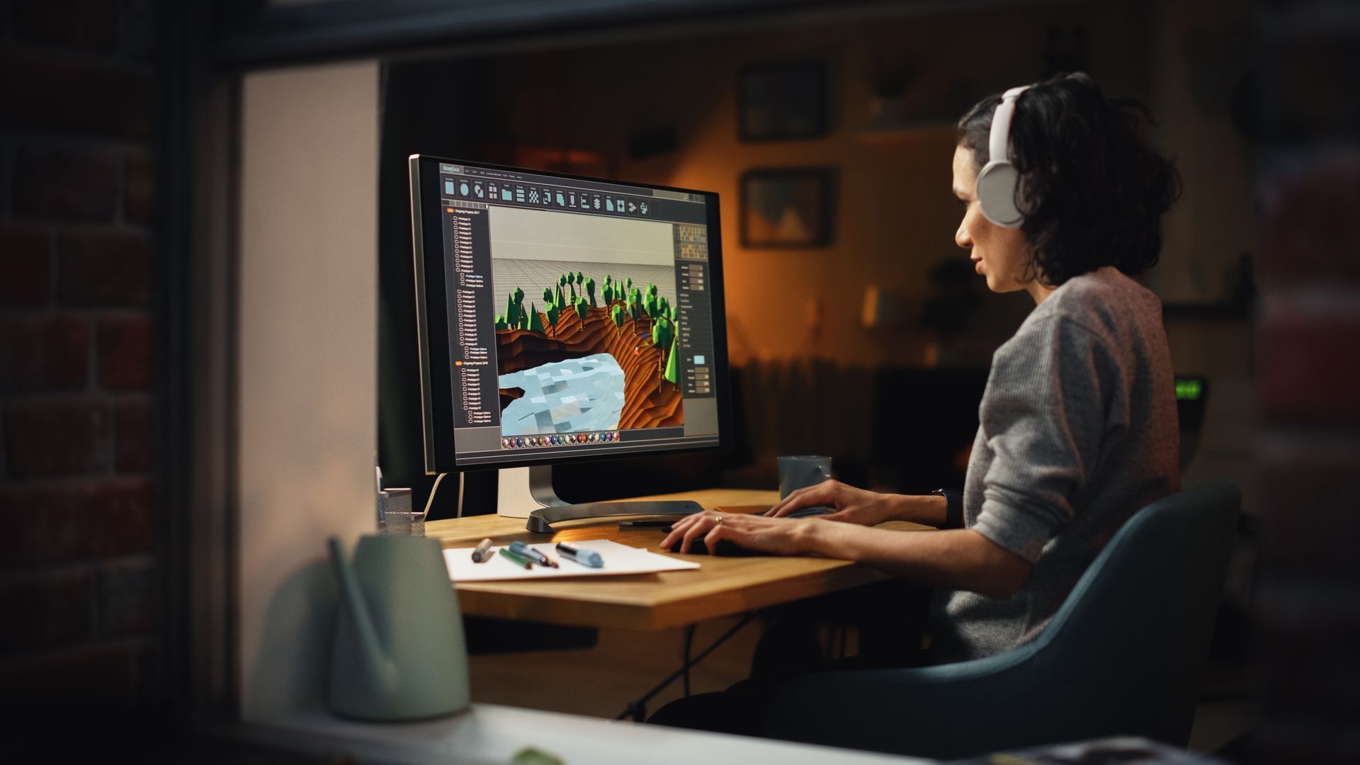 A person wearing headphones works at a desk on a computer, editing a digital landscape image in a dimly lit, cozy room.