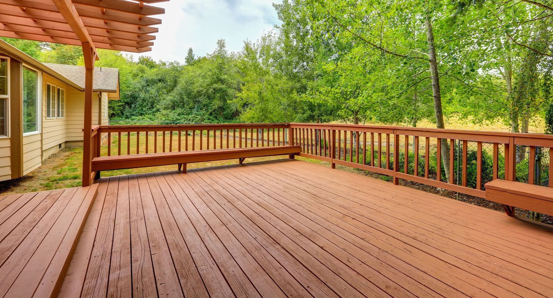 A spacious, reddish-brown wooden deck with built-in benches, a pergola section, and a railing overlooking a grassy yard.