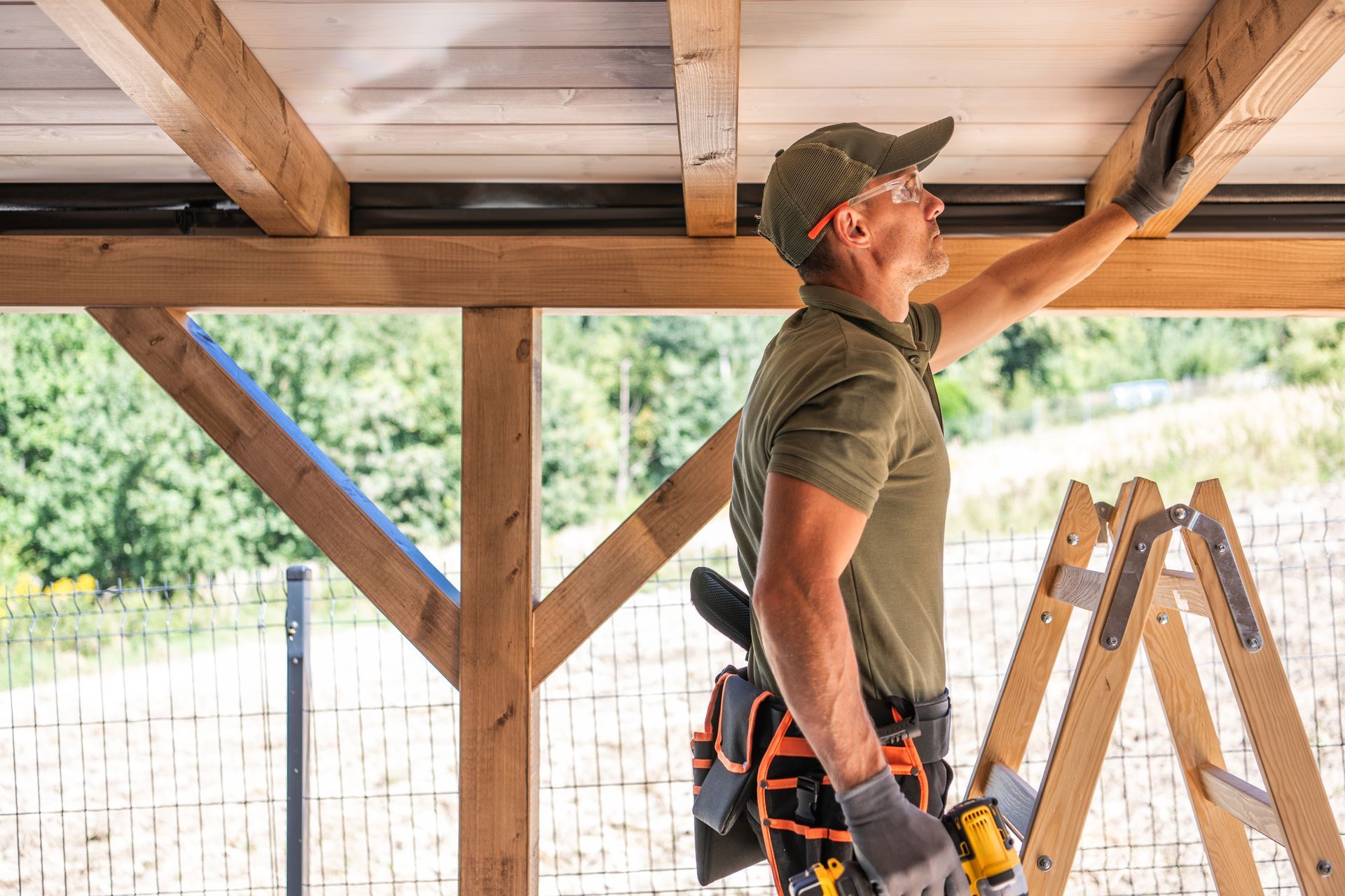 A person in a utility vest and cap reaches up to inspect a wooden ceiling structure while standing on a ladder outdoors.
