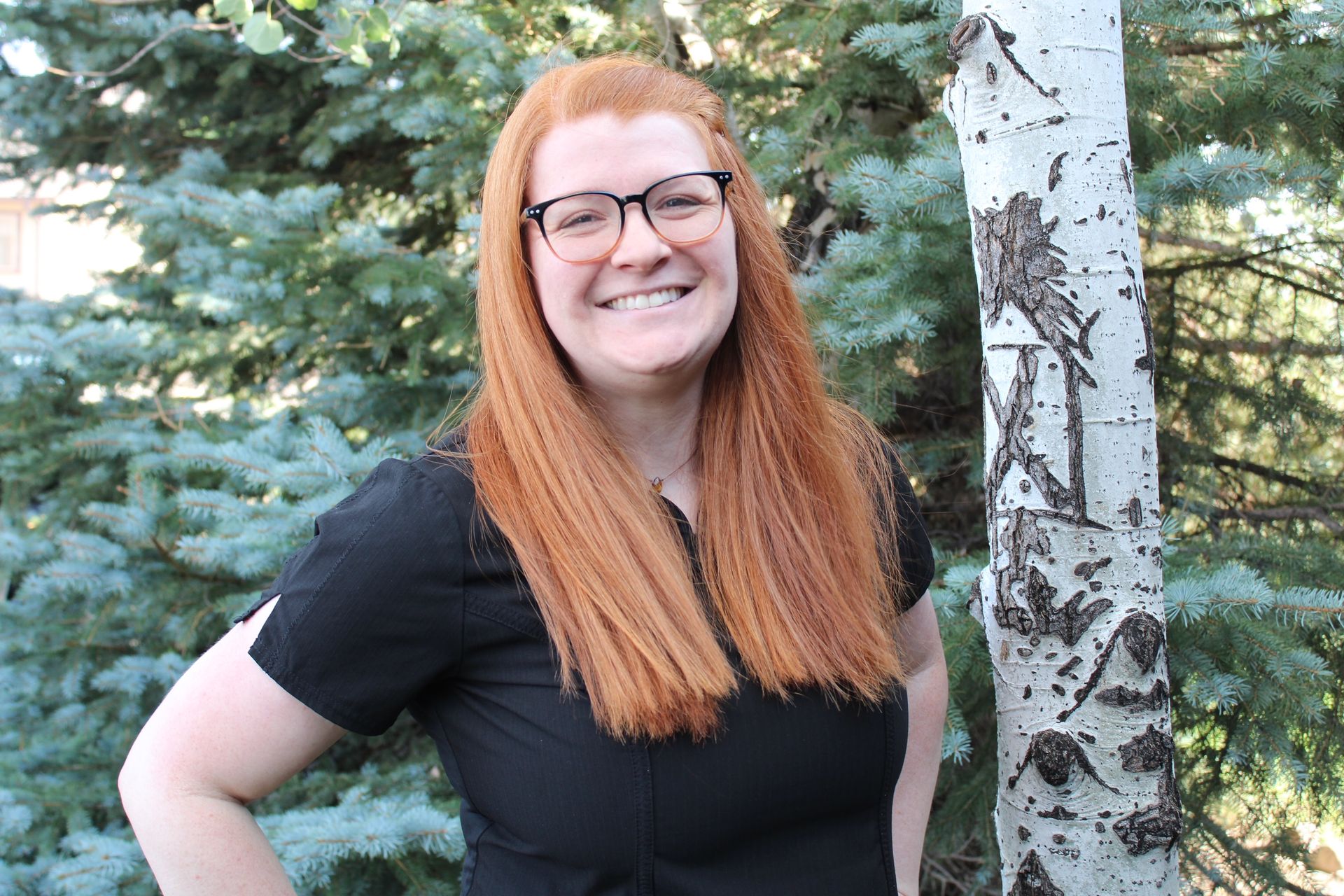 A woman with red hair and glasses is standing in front of a tree.