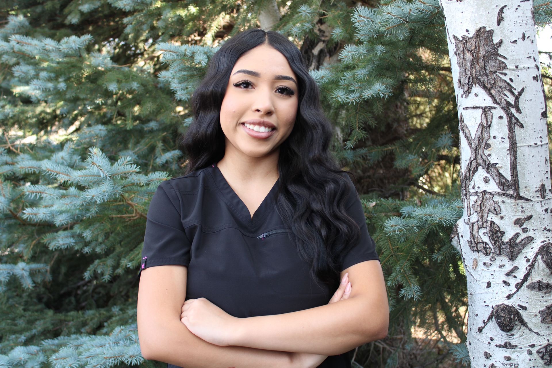 A woman in a black scrub top is standing in front of a tree with her arms crossed.