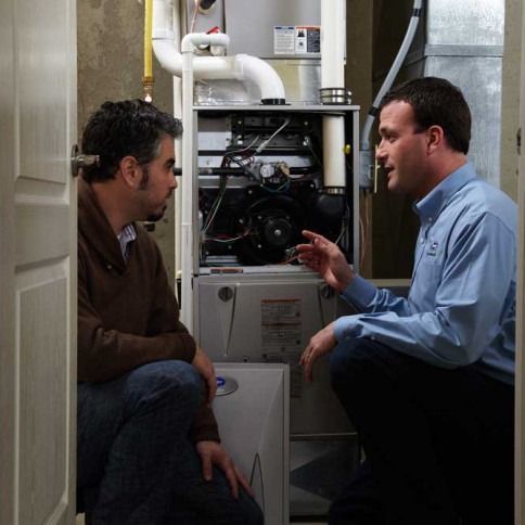 Two men examine a furnace. One kneels, pointing; the other watches. Inside a utility room.