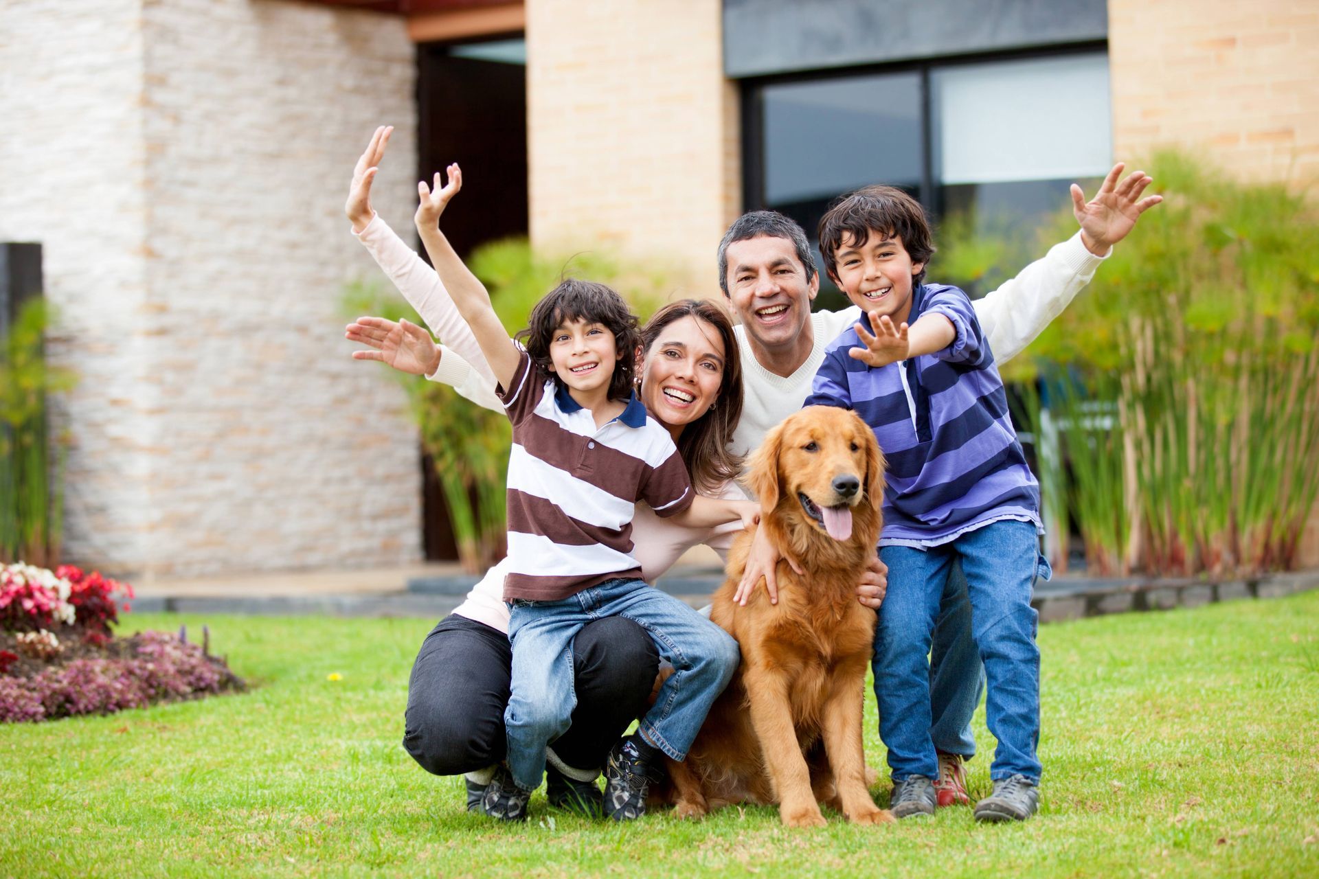 Family smiling and waving while sitting with a golden retriever on a lawn outside a house