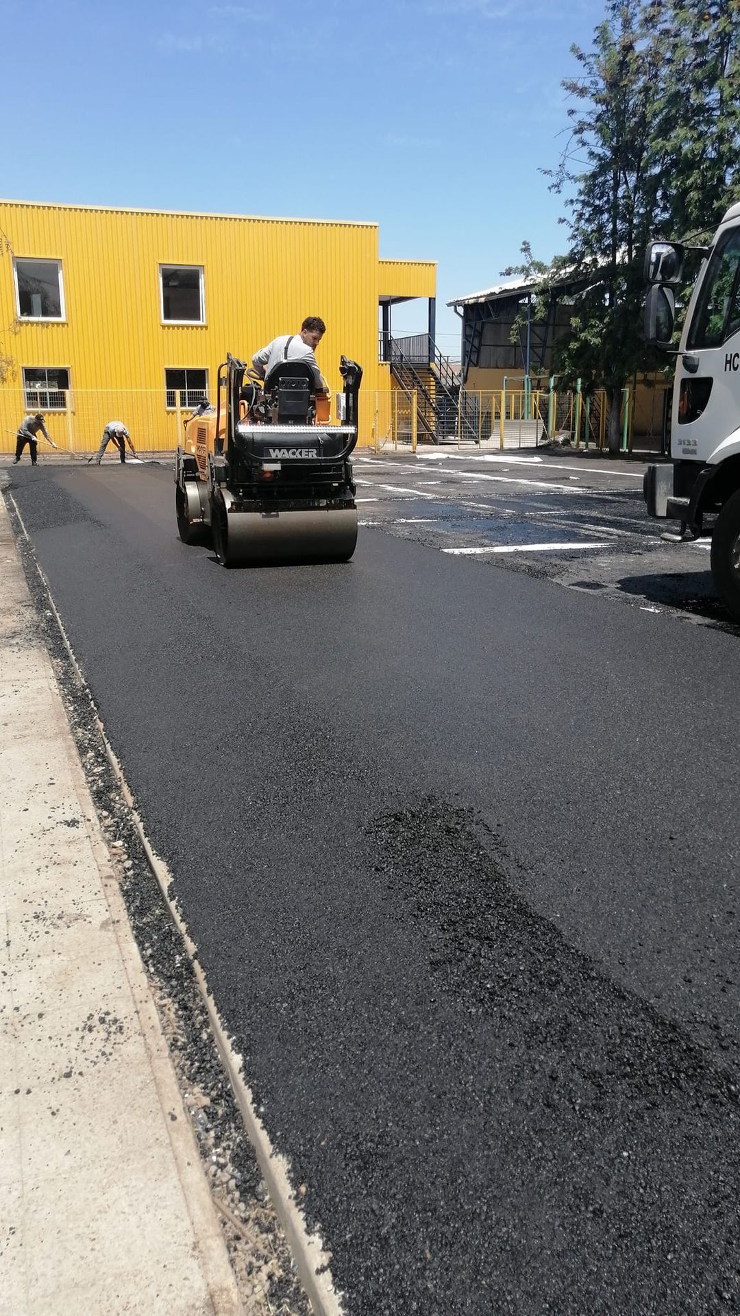Asfaltos Patricio Contreras - Un hombre conduce un rodillo por una carretera delante de un edificio amarillo.