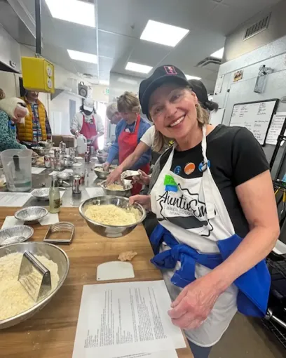 A woman in an apron is smiling in a kitchen while preparing food.