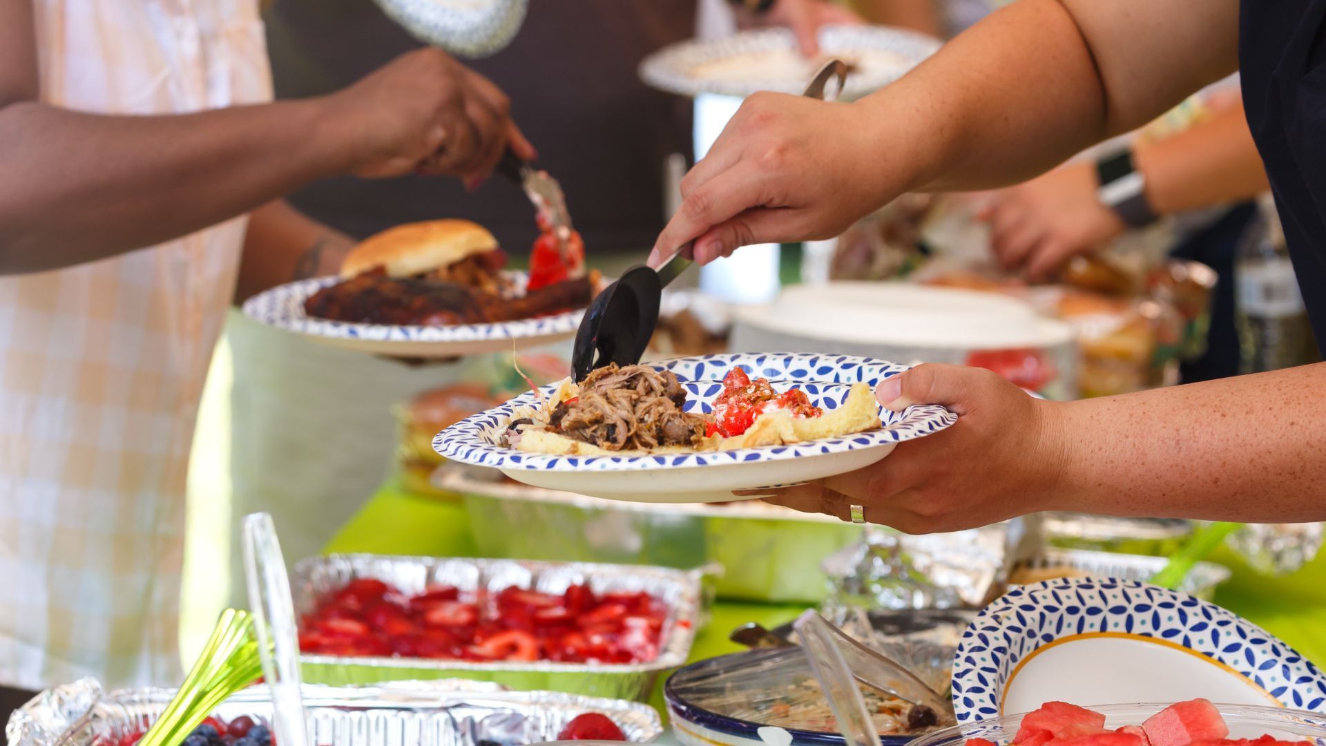 Close-up of a community member serving pulled pork at a buffet-style picnic with fresh watermelon on the table.