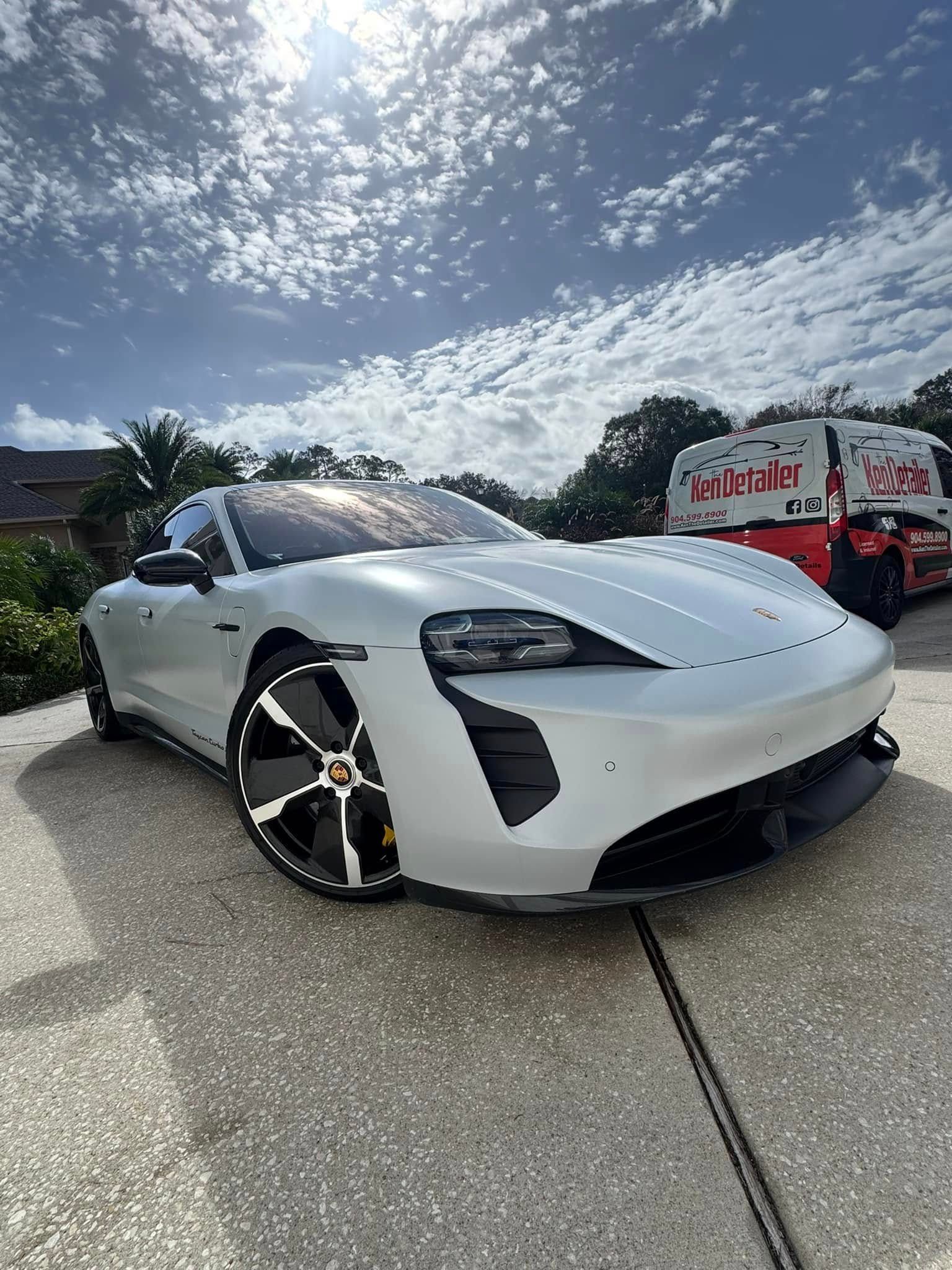 Silver Porsche Taycan parked in driveway with a red and white truck in the background, under a bright sky.
