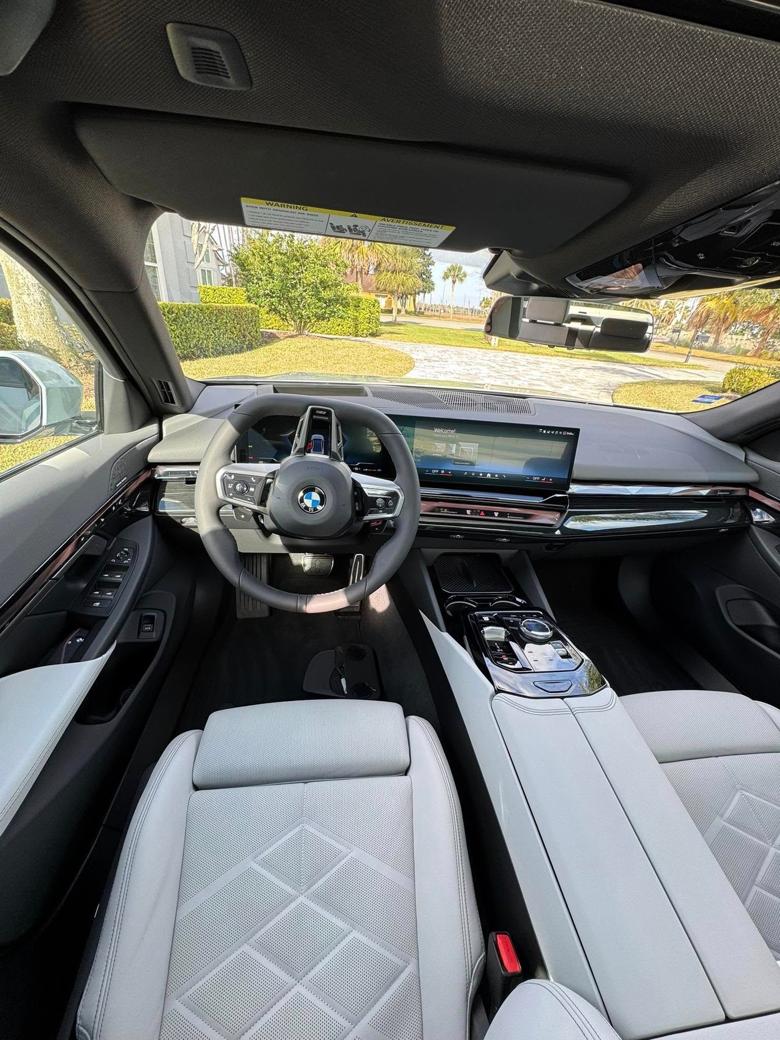 Interior view of a modern BMW car, showing the dashboard, steering wheel, and white seats with a view of the outside.