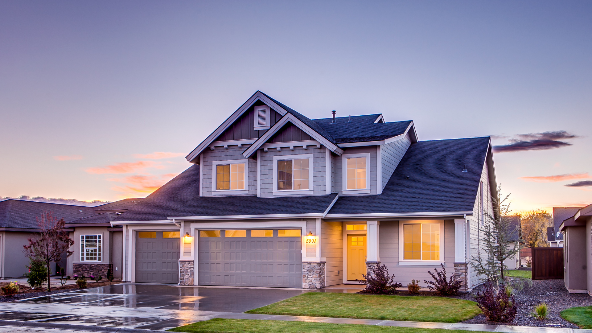 Suburban two-story house with lit windows at sunset, garage doors closed, and wet driveway