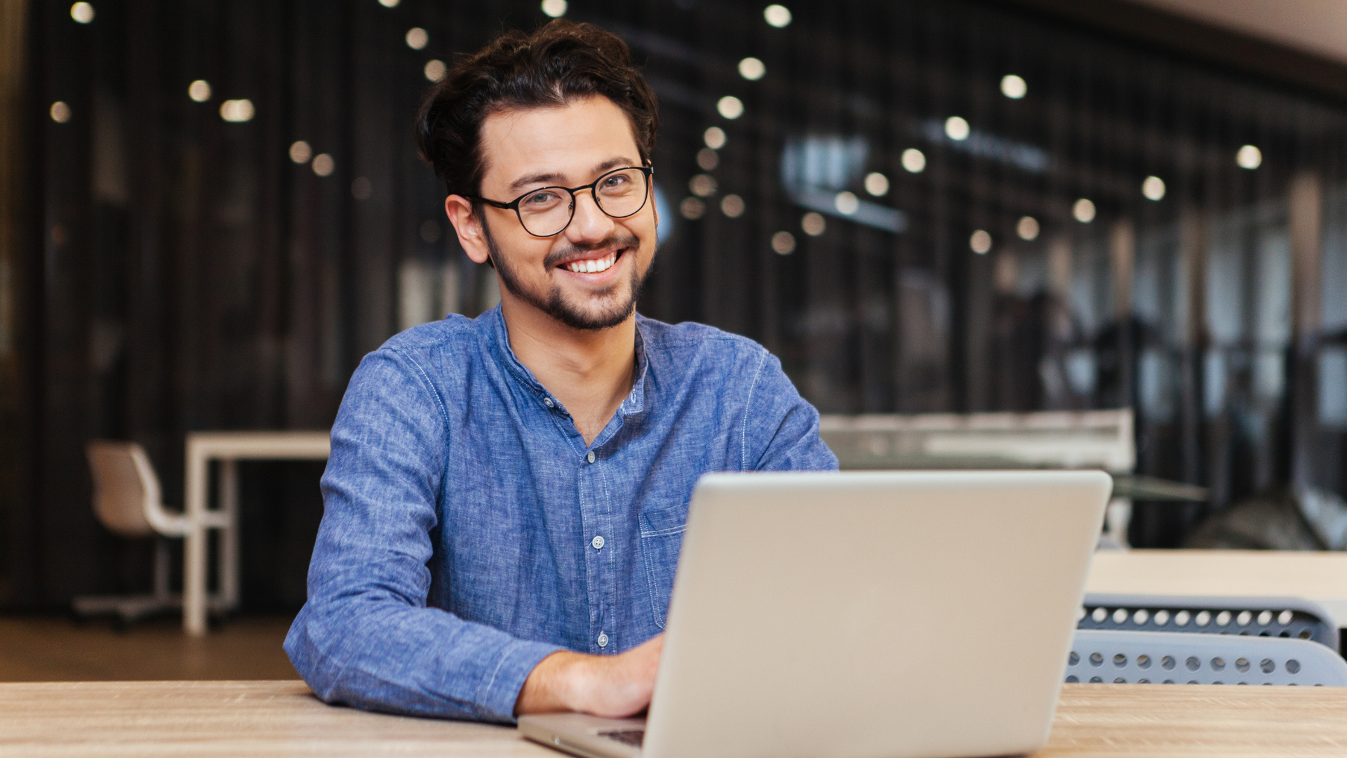 Smiling man in a blue shirt using a laptop at a table indoors