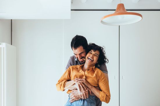 Man embracing and laughing with a woman; they are indoors with a modern light fixture.