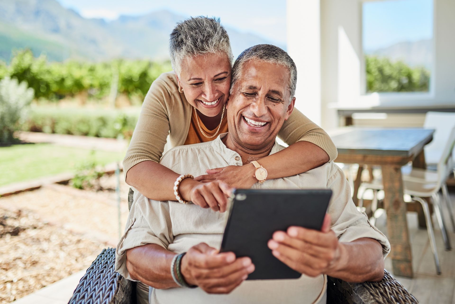 Elderly couple, smiling, looking at tablet outside. Woman hugs man from behind. Beige, natural light.