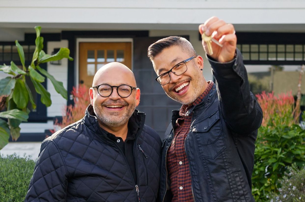 Two men smiling in front of a house; one holds up a key.