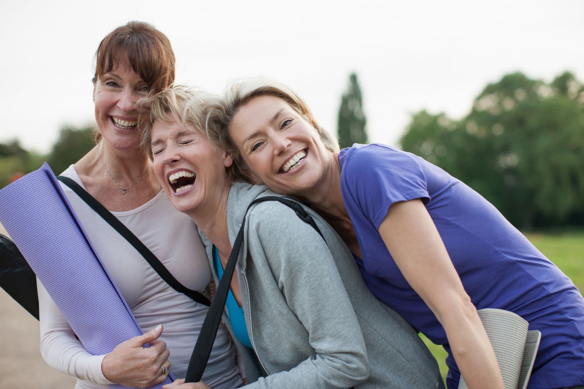 Three smiling women in casual wear outdoors, embracing, one holding a yoga mat.