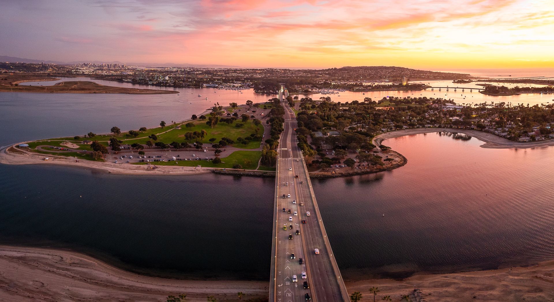 Bridge over water at sunset; cars driving, land with trees on either side, pink and orange sky.