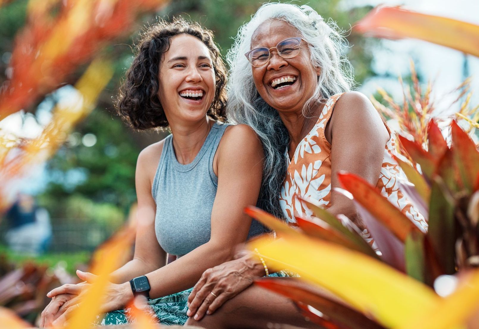 Two women laughing outdoors, one with gray hair, surrounded by colorful foliage.