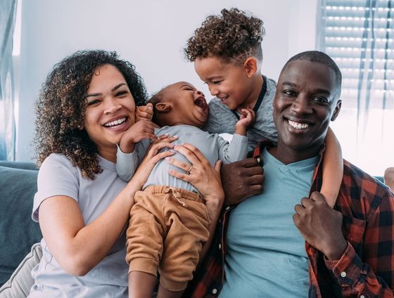 Happy family of four on a couch, smiling and laughing. Two children are held by their parents.