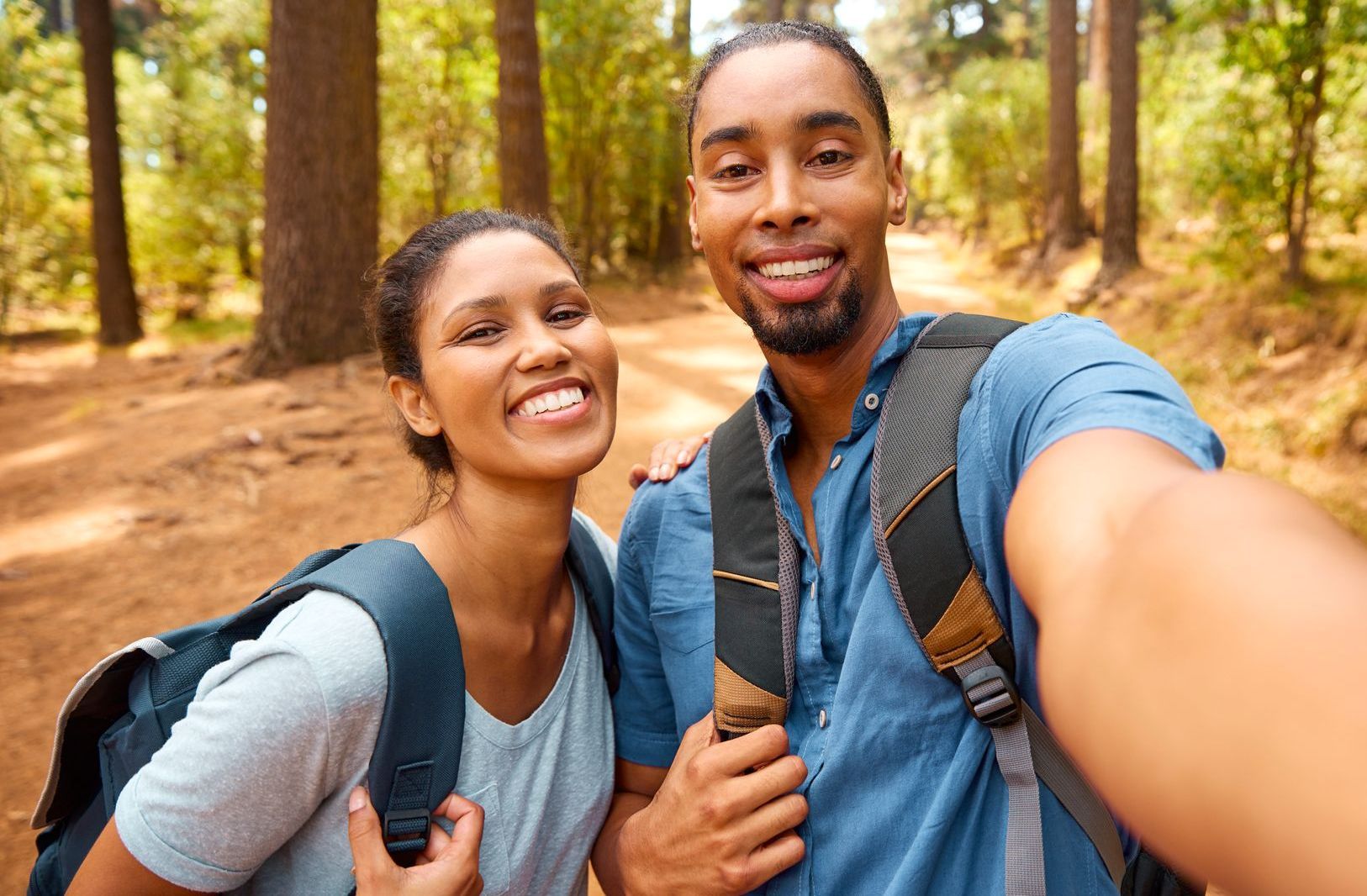 Couple with backpacks smiling, taking a selfie on a wooded trail.