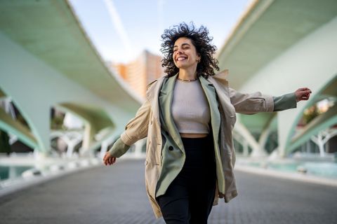 Woman with curly hair smiles and runs on a bridge. She wears a trench coat and black pants.