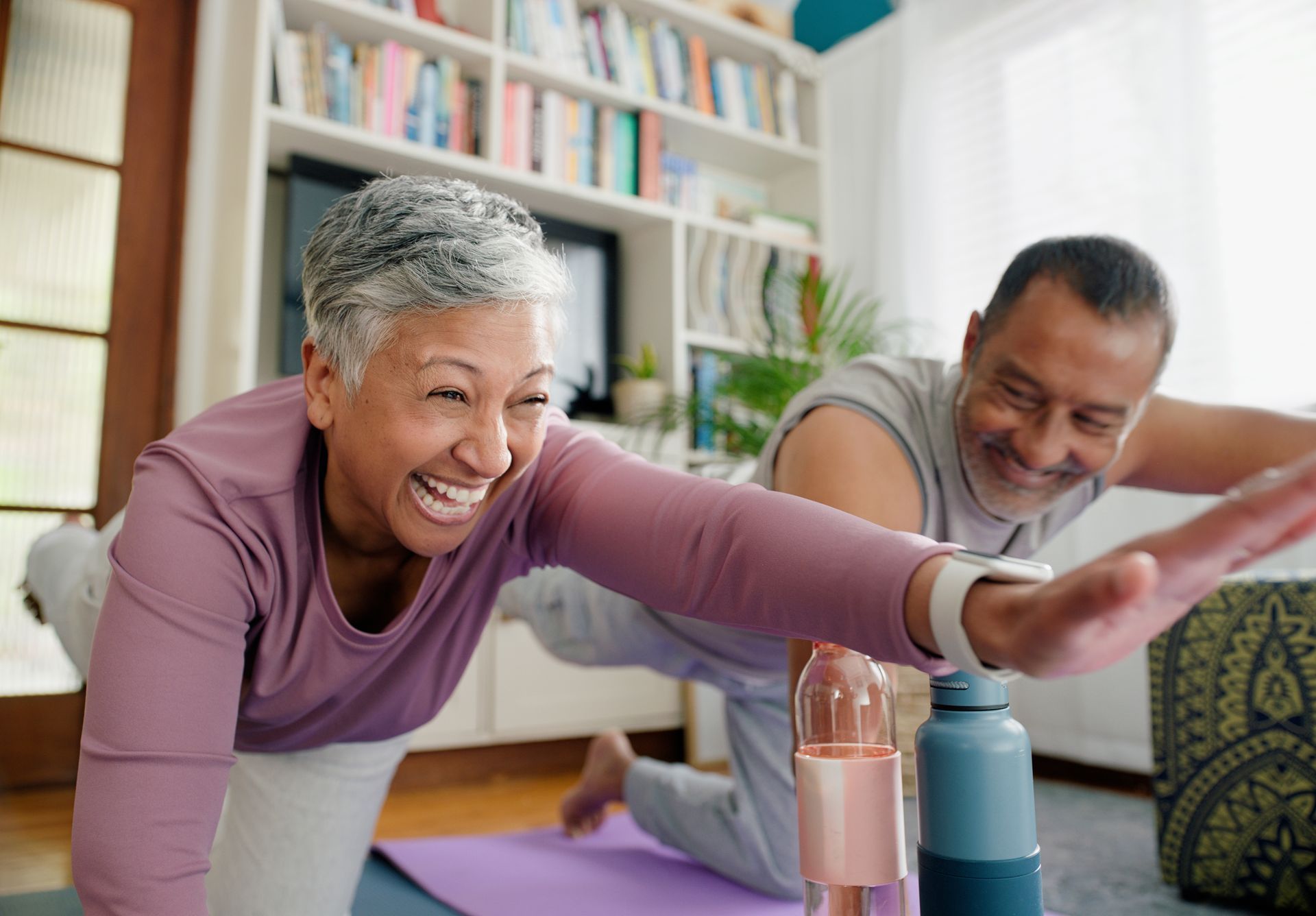 Smiling older couple exercising together on a yoga mat in a home setting.