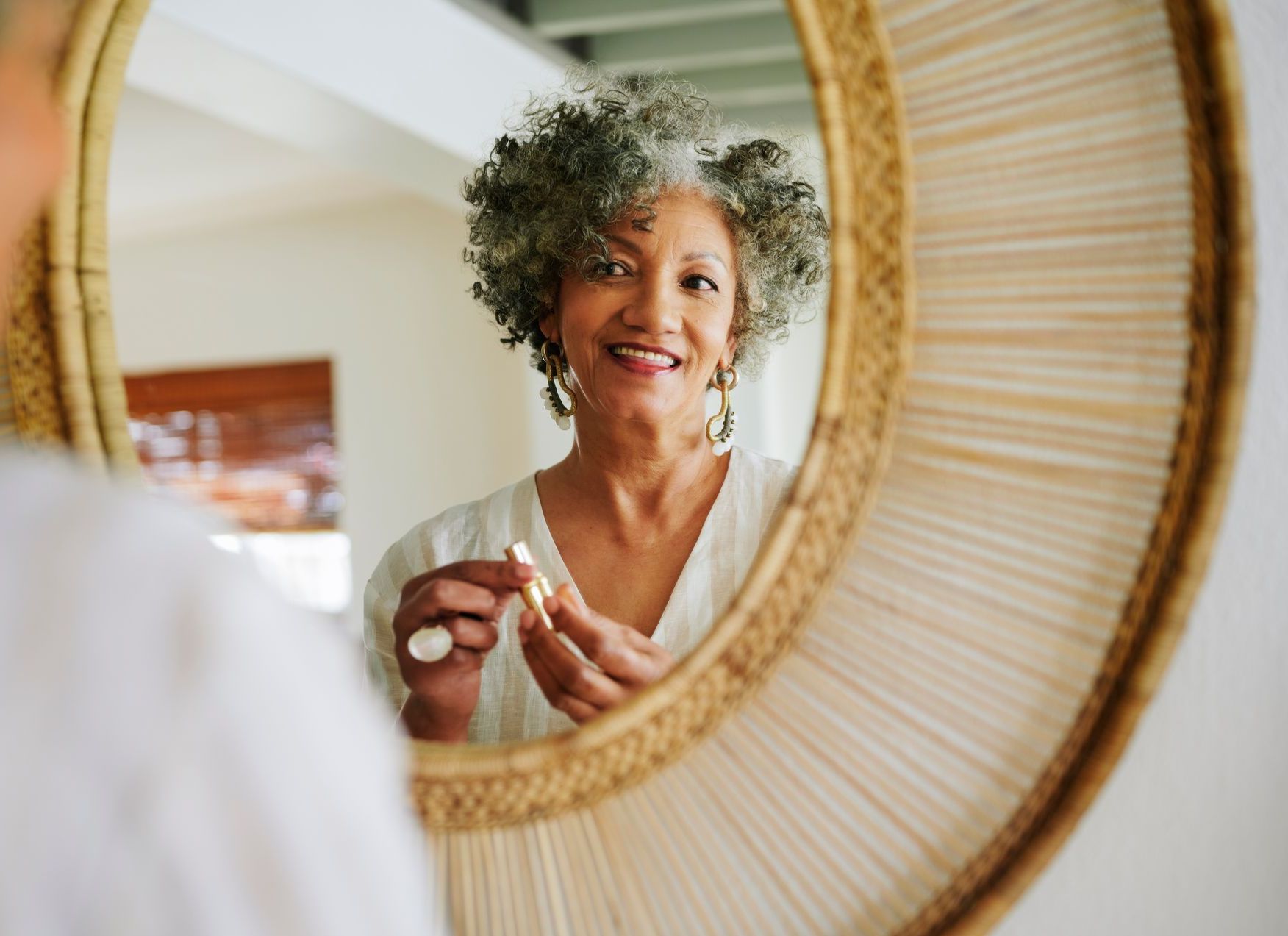 Woman with gray curly hair smiles in mirror, holding lipstick.